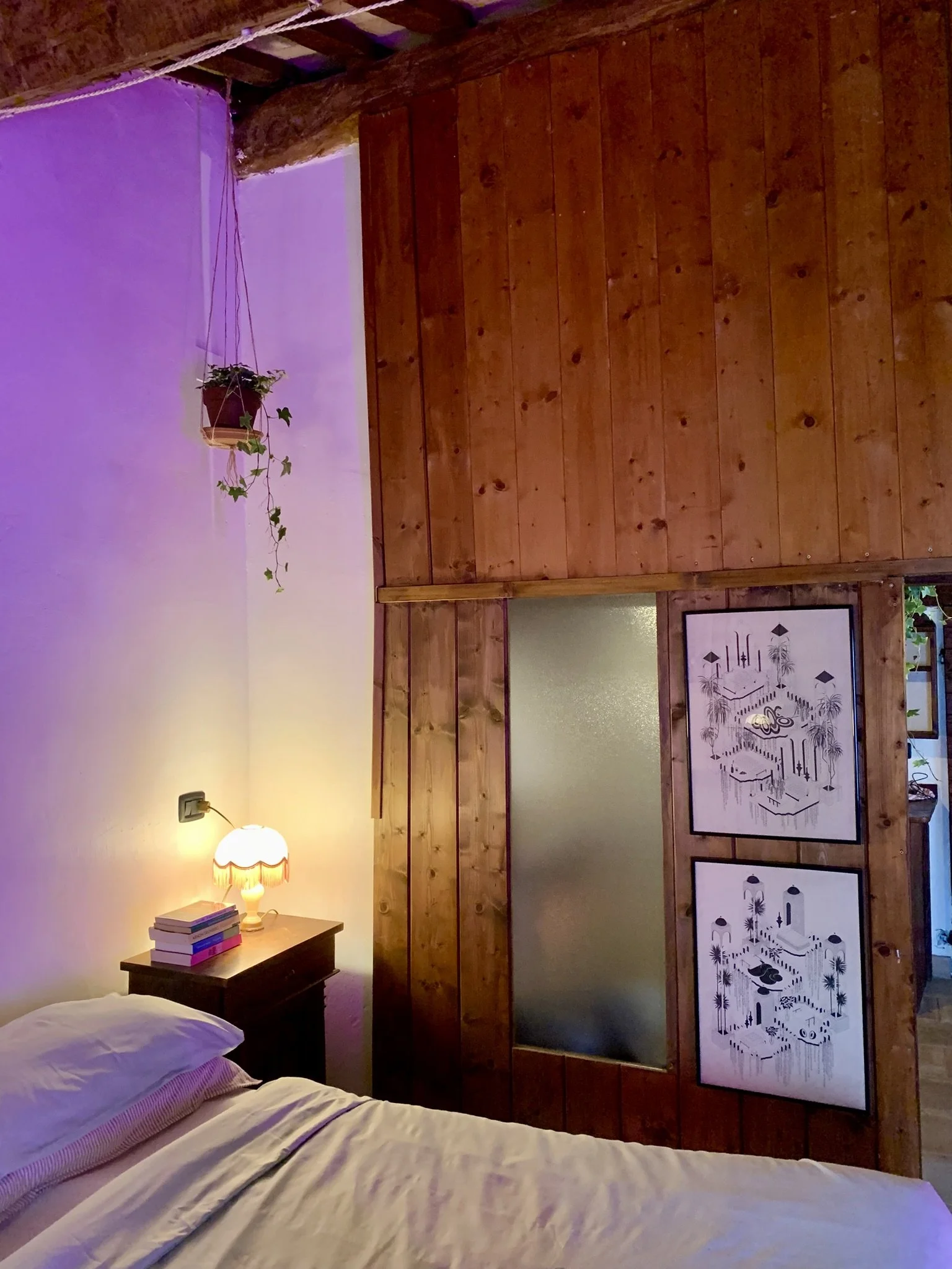 Bedroom with a bed in the foreground, a nightstand with several books and a lamp, purple lighting on the wall, wooden wall paneling, and framed artwork.