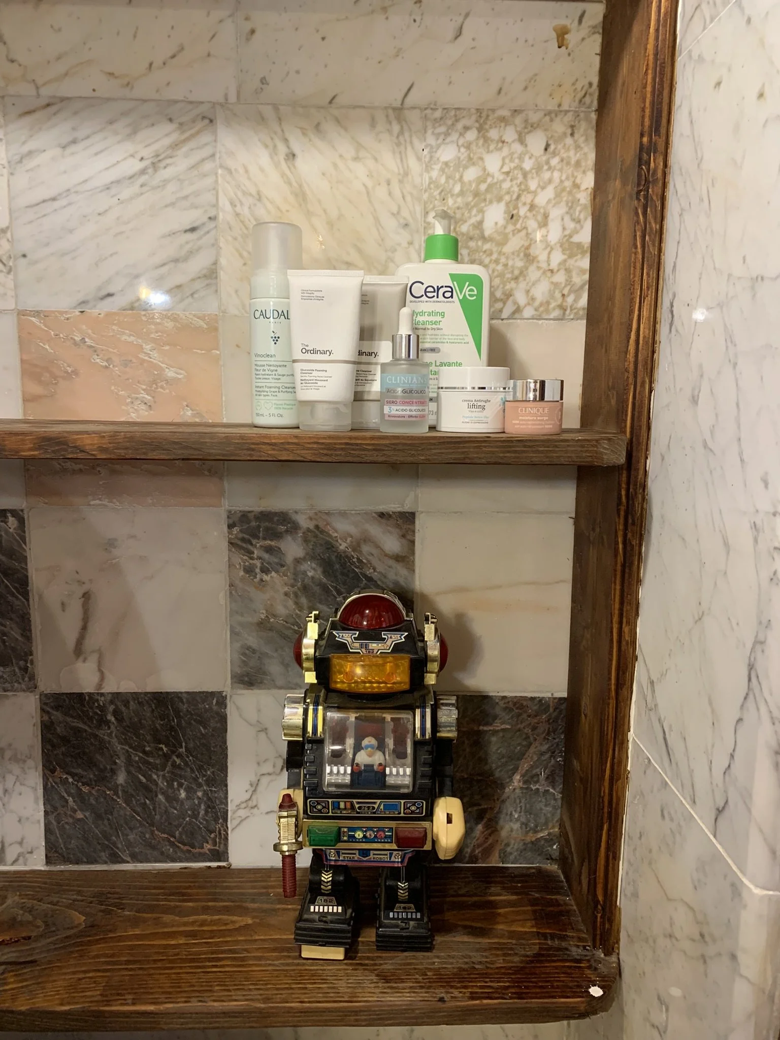 A marble tile bathroom wall with a wooden shelf holding skincare products and a vintage toy robot in front.