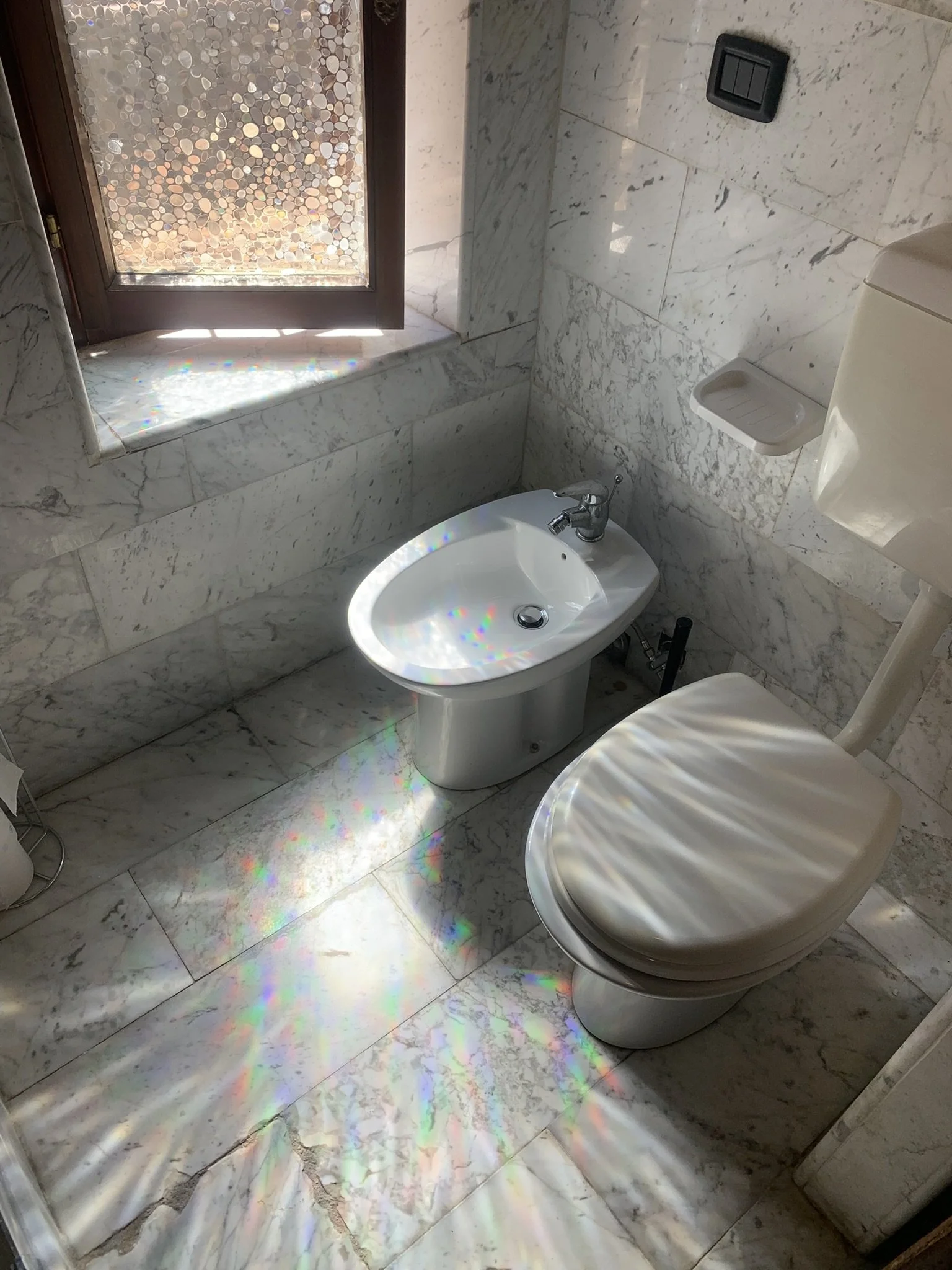A marble bathroom with a toilet, bidet, and window with a mosaic pattern. Sunlight creates a rainbow effect on the marble floor and wall.