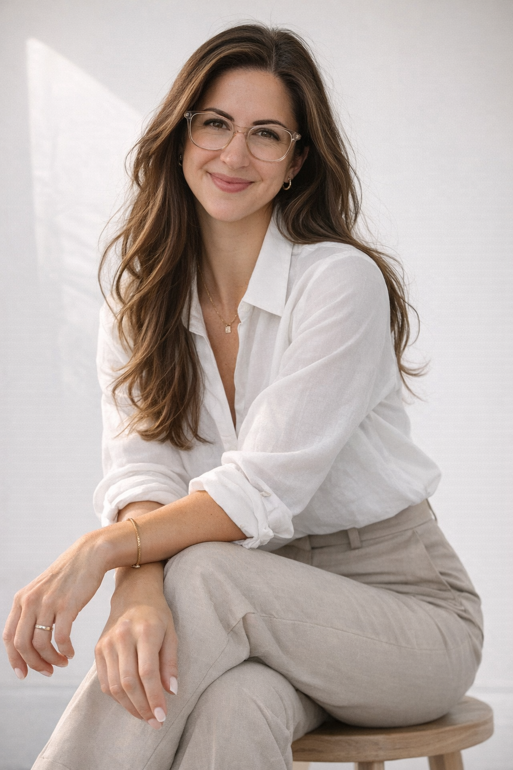 A young woman with long brown hair, wearing clear glasses, a white button-up shirt with sleeves rolled up, and beige pants, sitting on a wooden stool and smiling at the camera.