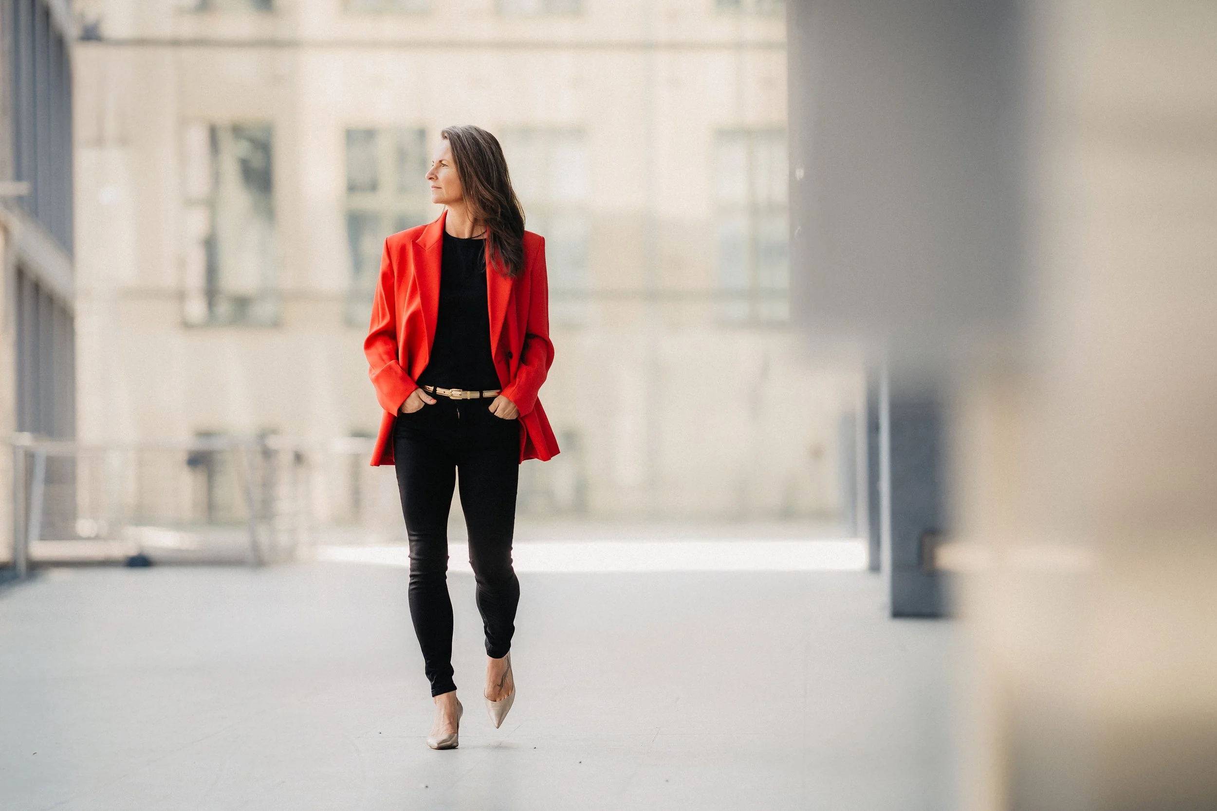 Frau in rotem Blazer, schwarzem Hemd und schwarzen Hosen geht auf einer modernen, urbanen Straße.