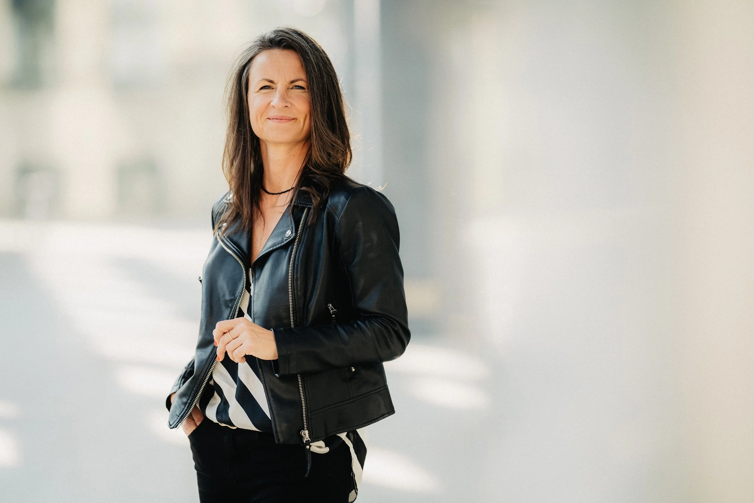 Eine Frau mit braunen Haaren trägt eine schwarze Lederjacke und steht vor einem hellen, unscharfen Hintergrund. Sie schaut einen Moment in die Kamera und lächelt leicht.