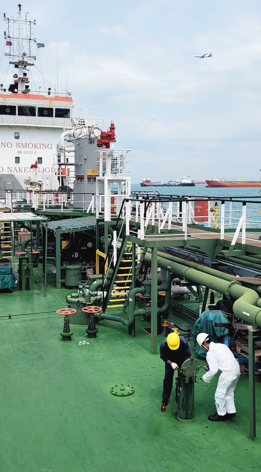 Two workers in safety gear inspecting equipment on the deck of a large industrial ship at sea with other ships and an airplane flying overhead.