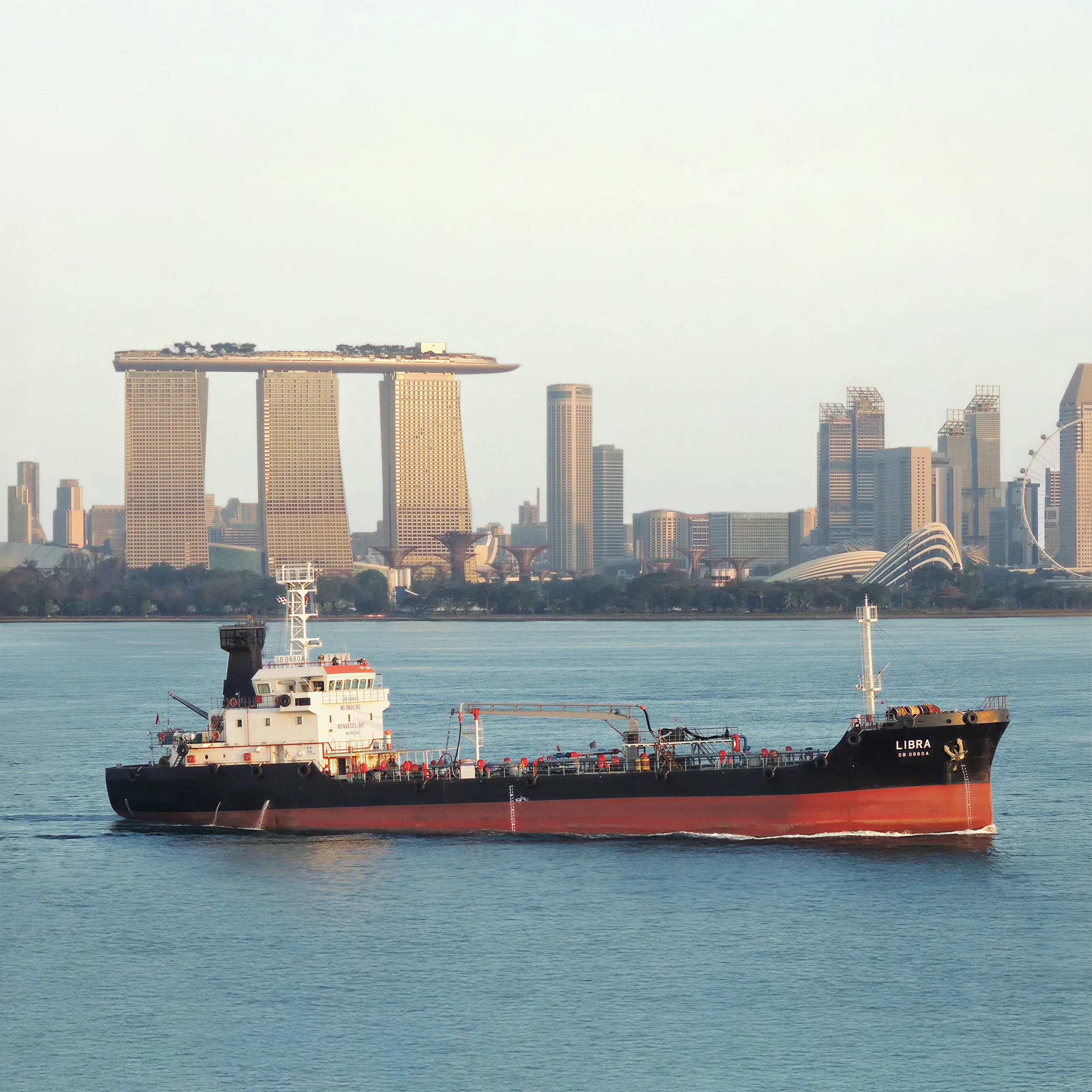 A ship named LIBRA sailing on a body of water with a city skyline featuring tall buildings and landmarks, including the Marina Bay Sands hotel, in the background.