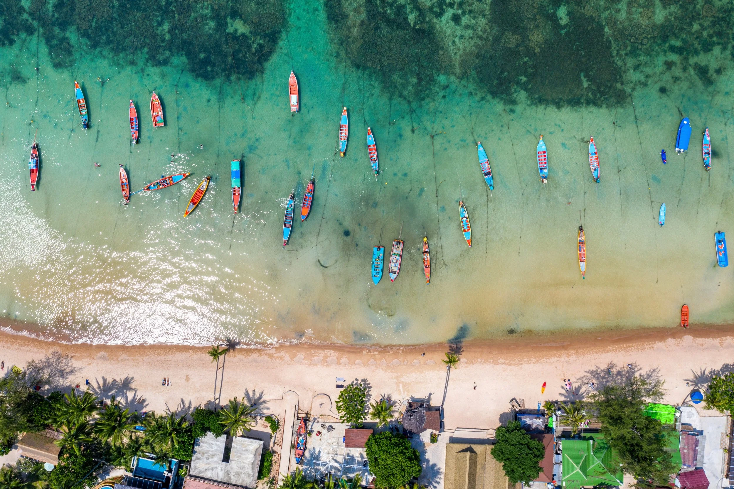 Aerial view of a tropical beach with boats anchored near the shore, palm trees, and beach houses.