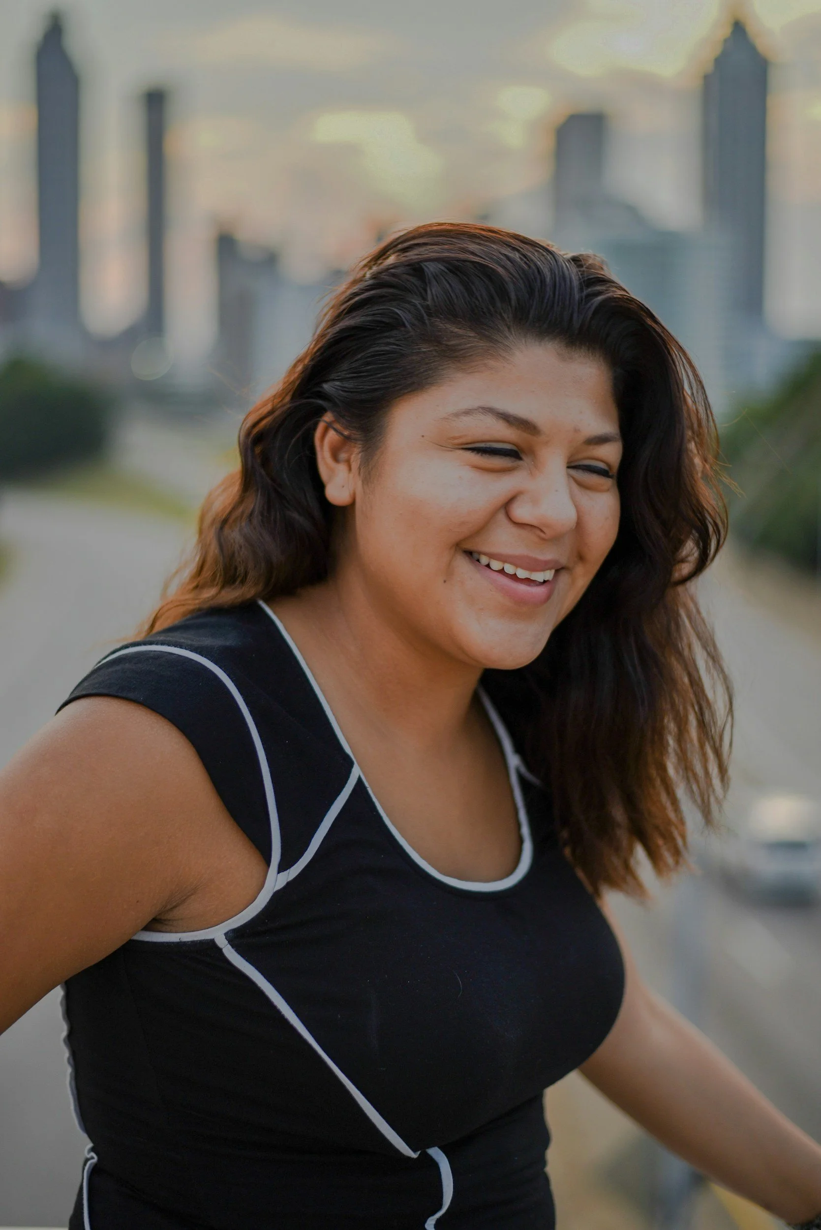 A smiling woman with dark, wavy hair outdoors with a city skyline in the background during sunset.