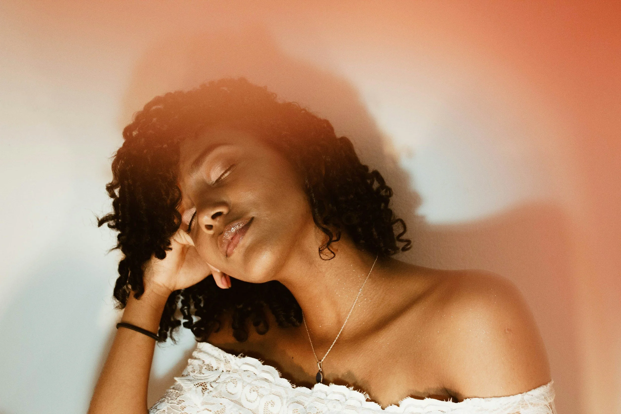 A woman with curly hair, wearing a white off-shoulder top, with eyes closed and a relaxed expression, touching her head with her left hand, against a plain background with a pinkish highlight.