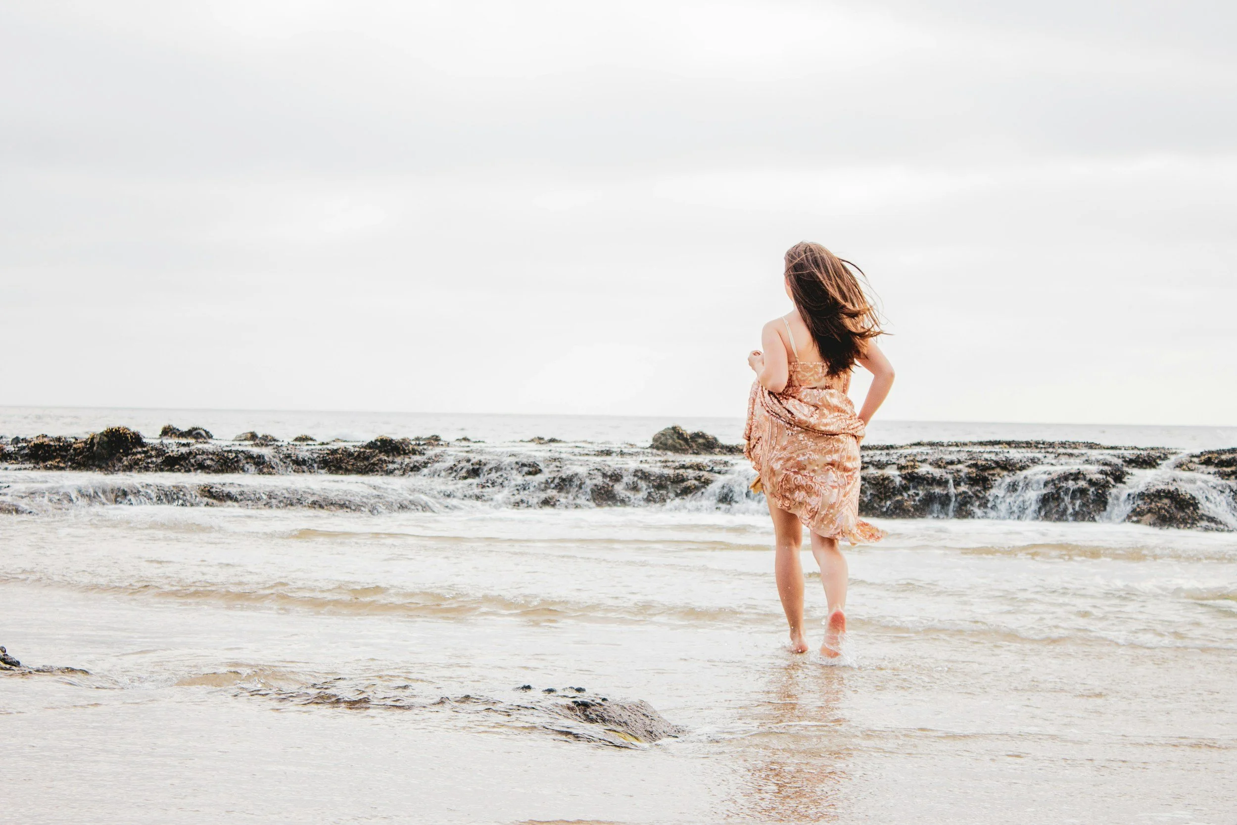 A woman in a beige dress walking on the beach near the shoreline with rocks and ocean waves, with her hair blowing in the wind under a cloudy sky.