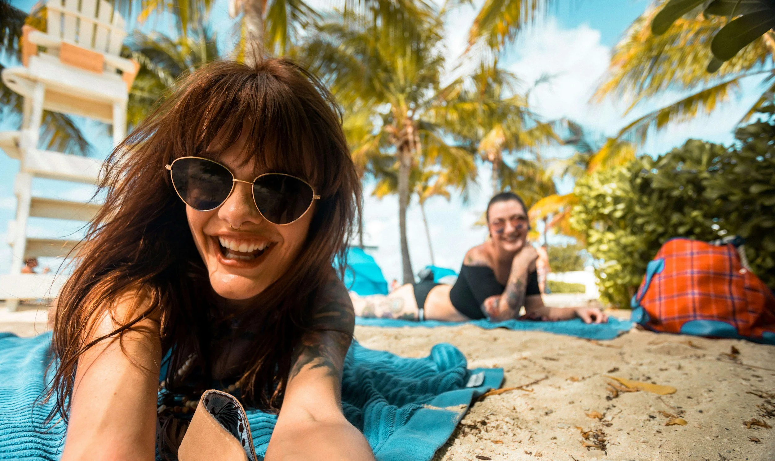 Two women lying on a beach with palm trees, smiling and laughing, sunny weather, one woman in the foreground wearing sunglasses and the other in the background with tattoos, beach bag and umbrellas nearby.