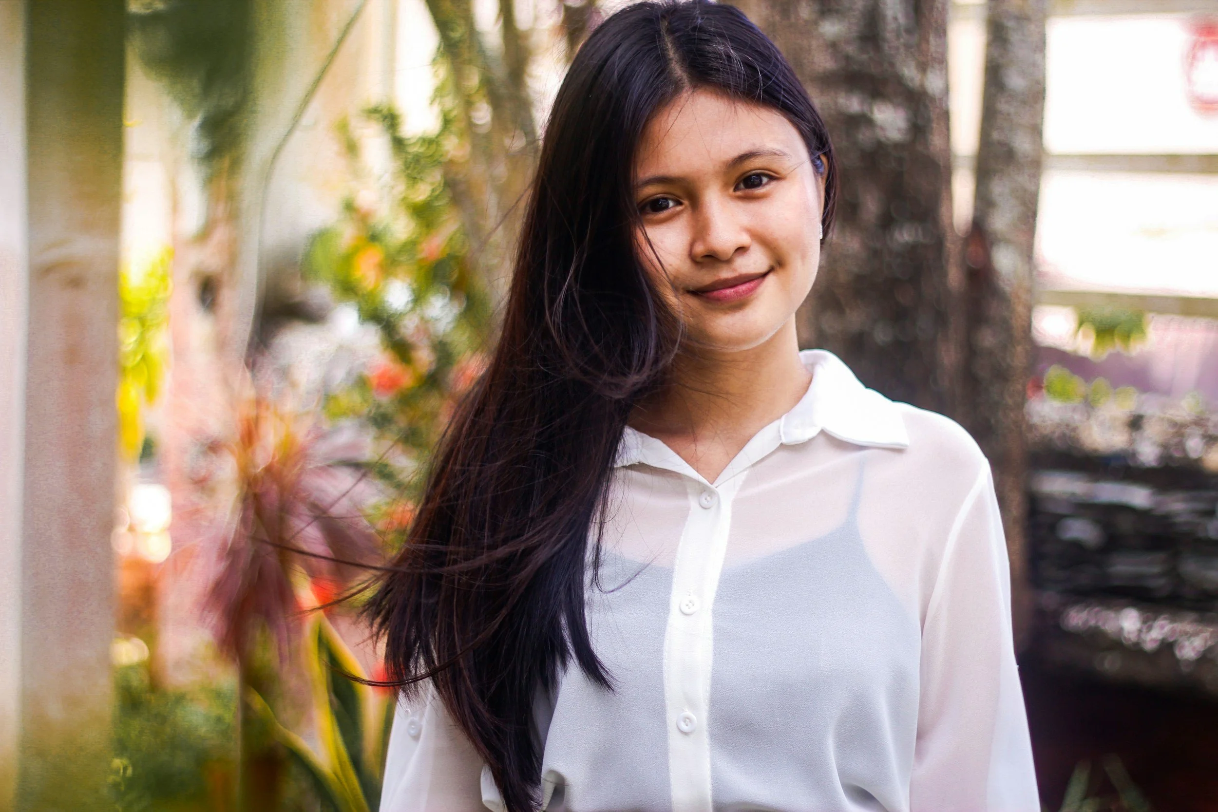 A young woman with long dark hair, wearing a white blouse, outdoors with trees in the background.