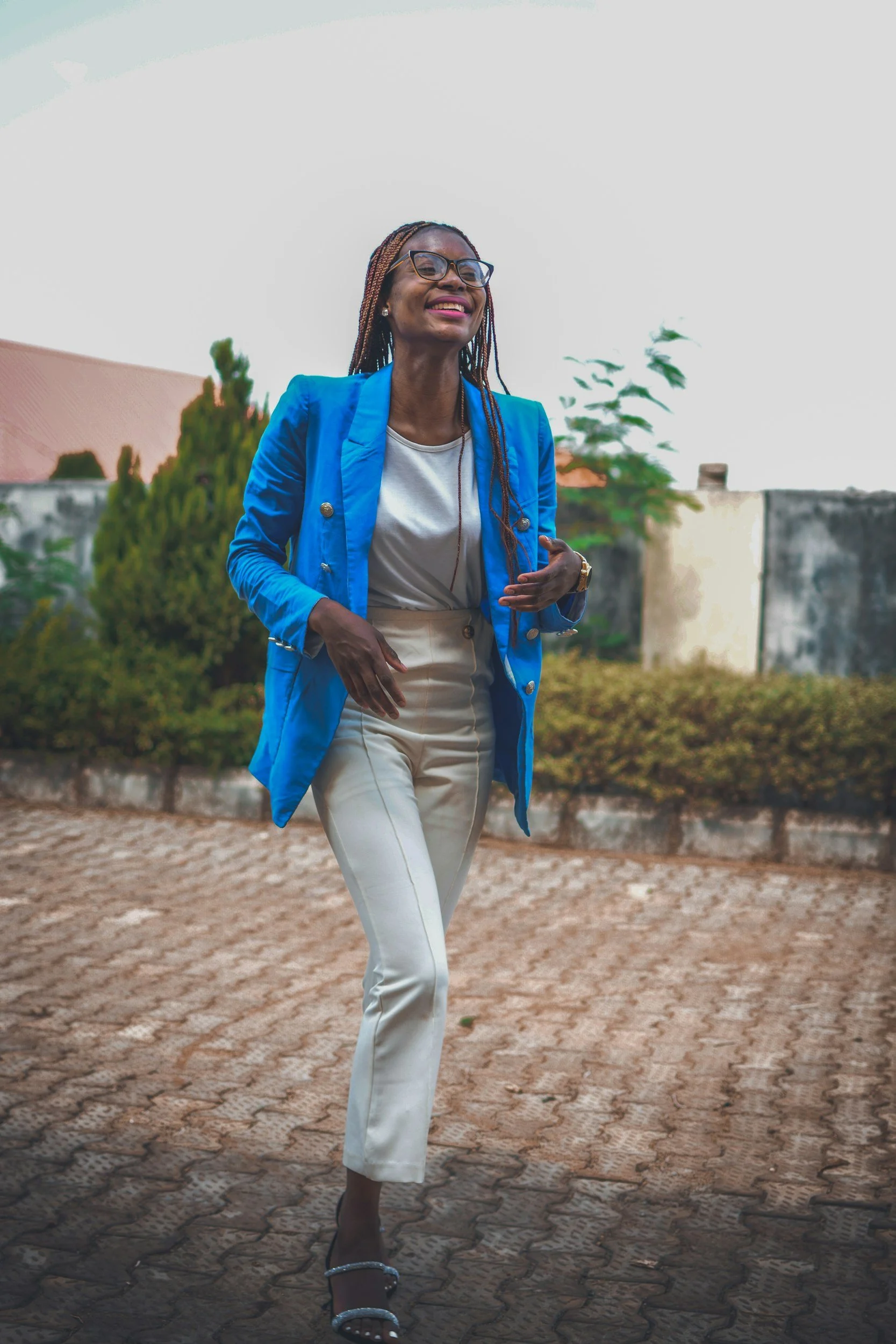 A woman with braided hair and glasses smiling, wearing a white shirt, blue blazer, white pants, and black heels, walking outdoors on a paved area with greenery and buildings in the background.
