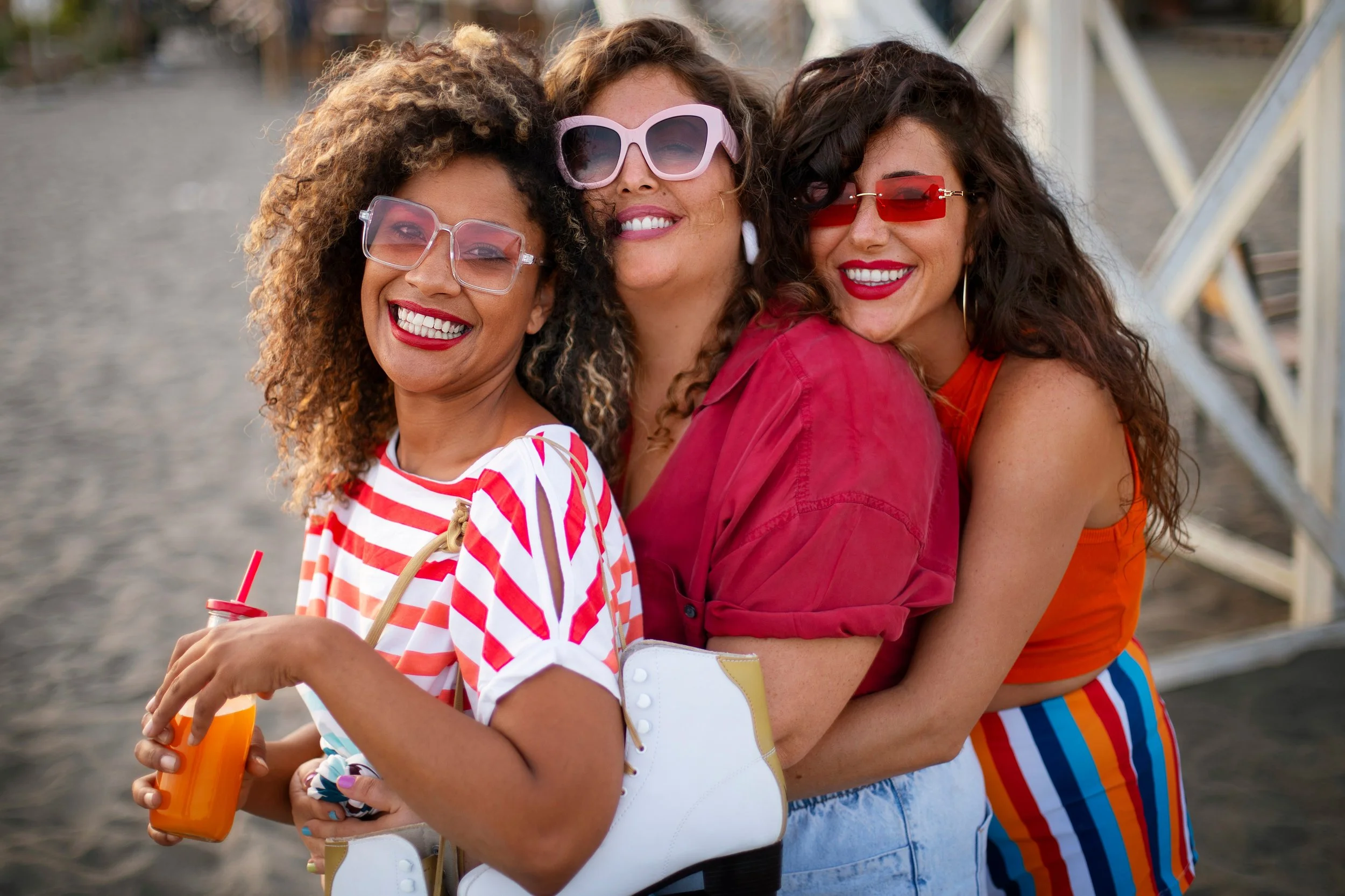 Three women with curly hair and sunglasses smiling, standing close together at a waterfront during daytime, wearing colorful outfits including stripes and solid colors.