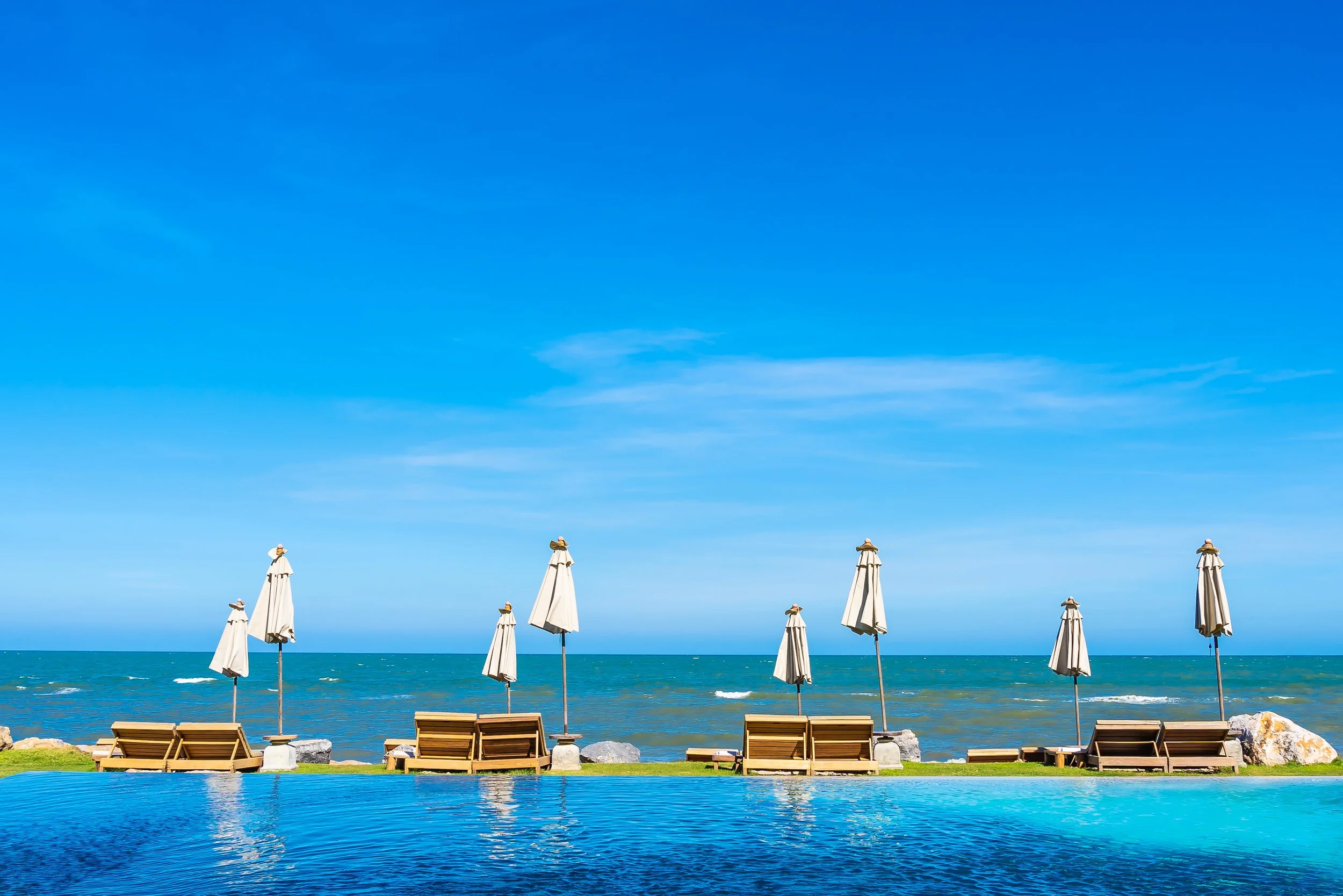 Beach scene with a row of lounge chairs and umbrellas facing the ocean, a clear blue sky overhead, and an infinity pool in the foreground.