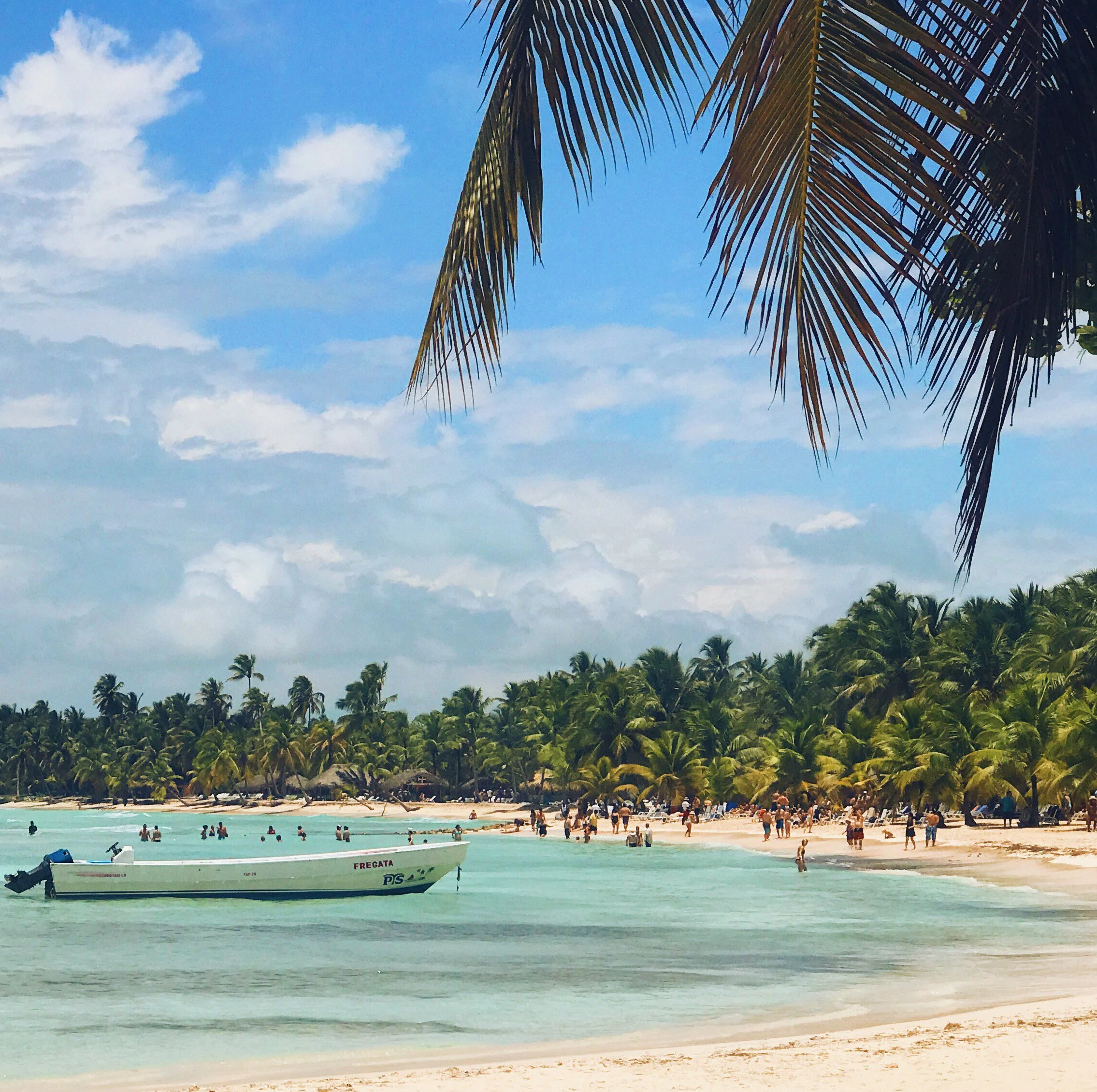 Tropical beach scene with palm trees, a boat labeled 'Fregata' in the water, and many people on the sandy shoreline and in the ocean under a partly cloudy sky.