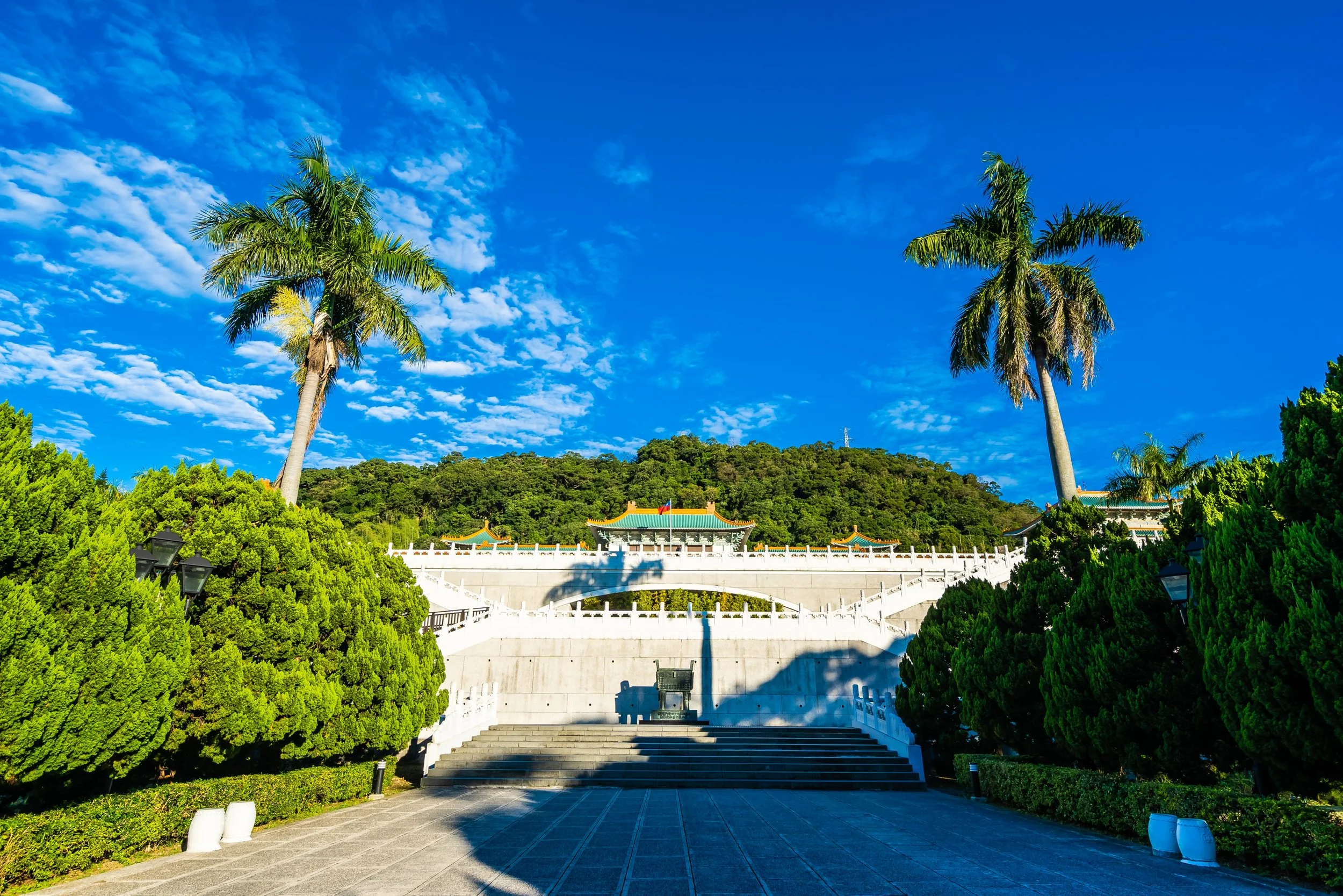 A scenic view of a park with two tall palm trees, bushes, and traditional Asian-style buildings with green and red roofs in the background, under a bright blue sky with scattered clouds.