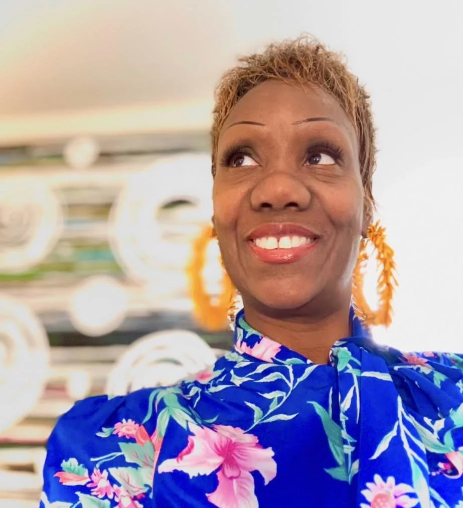A woman with short, light brown hair and orange earrings, smiling and looking to her left, dressed in a blue floral top.