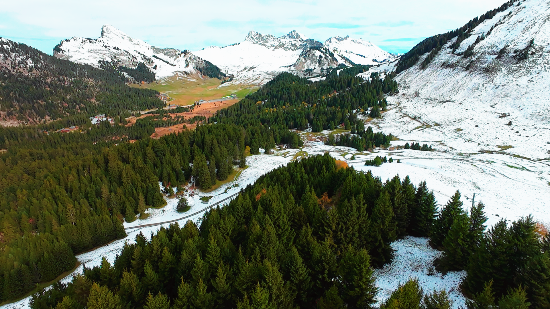 Paysage de montagnes enneigées, forêt dense, vallée, et petites maisons dans un environnement alpin.