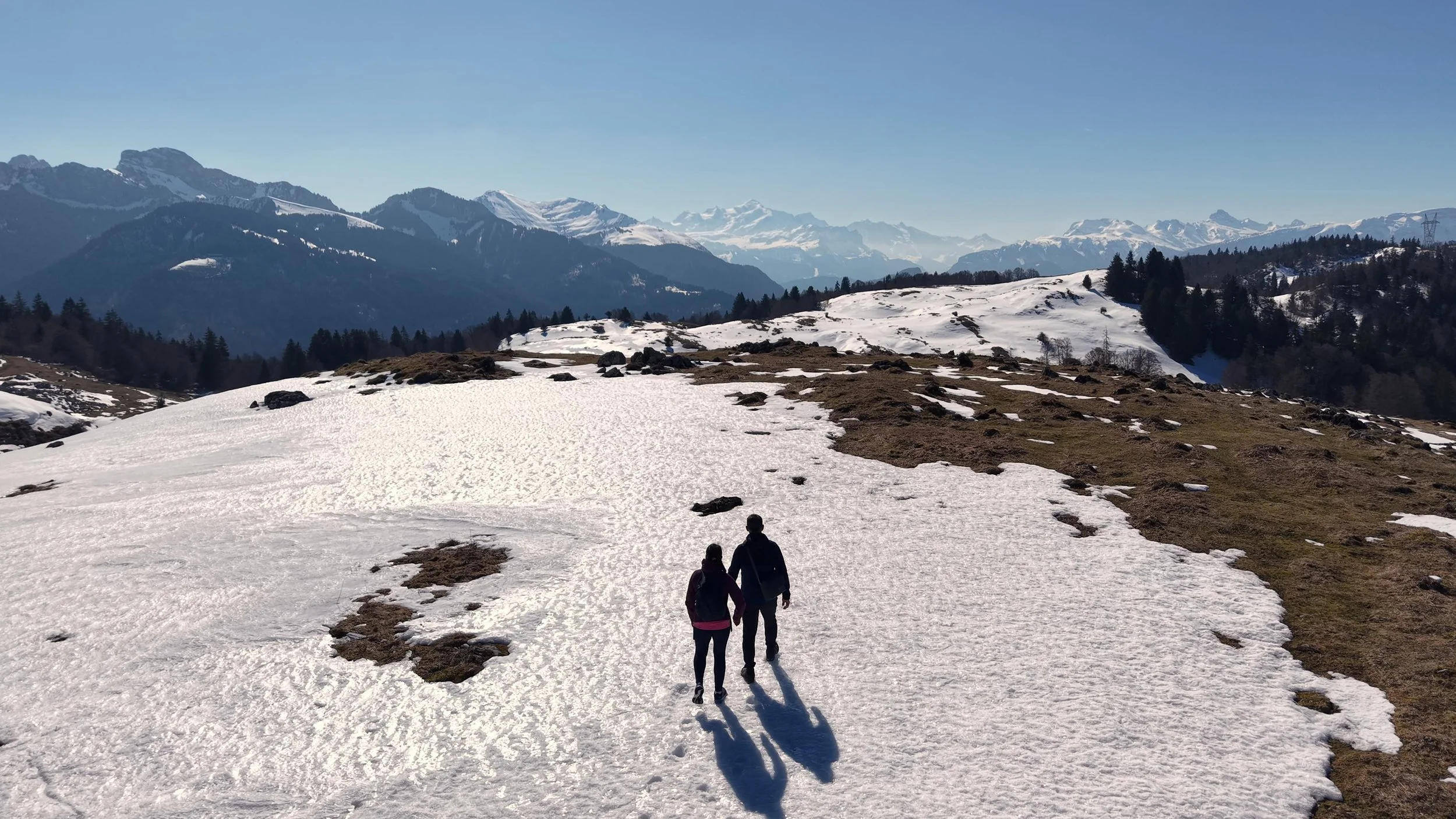 Deux personnes marchent sur un chemin enneigé dans un paysage de montagne avec des sommets enneigés en arrière-plan et des arbres autour.