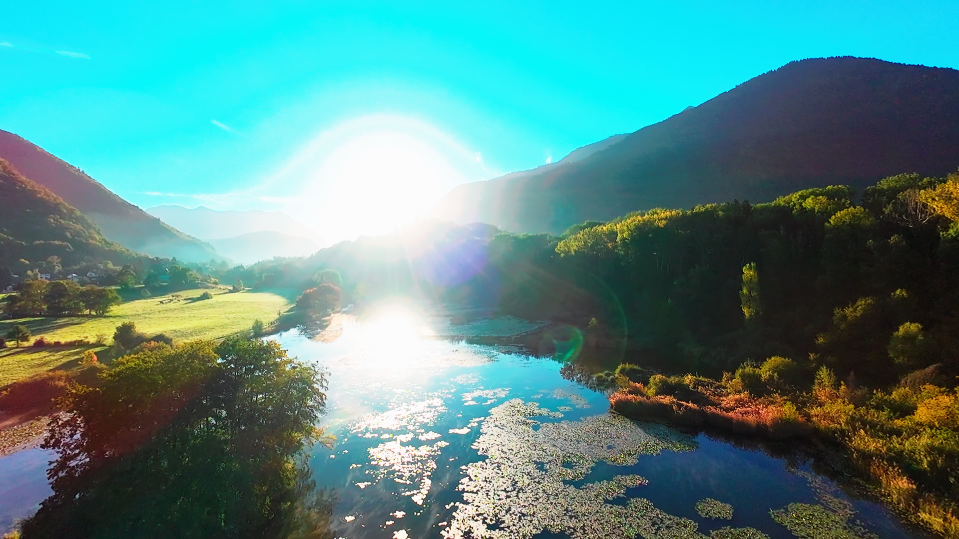 Un paysage de montagne avec un lac au premier plan, la lumière du soleil brillant à l'horizon, créant des réflexions sur l'eau, entouré d'arbres et de collines verdoyantes.
