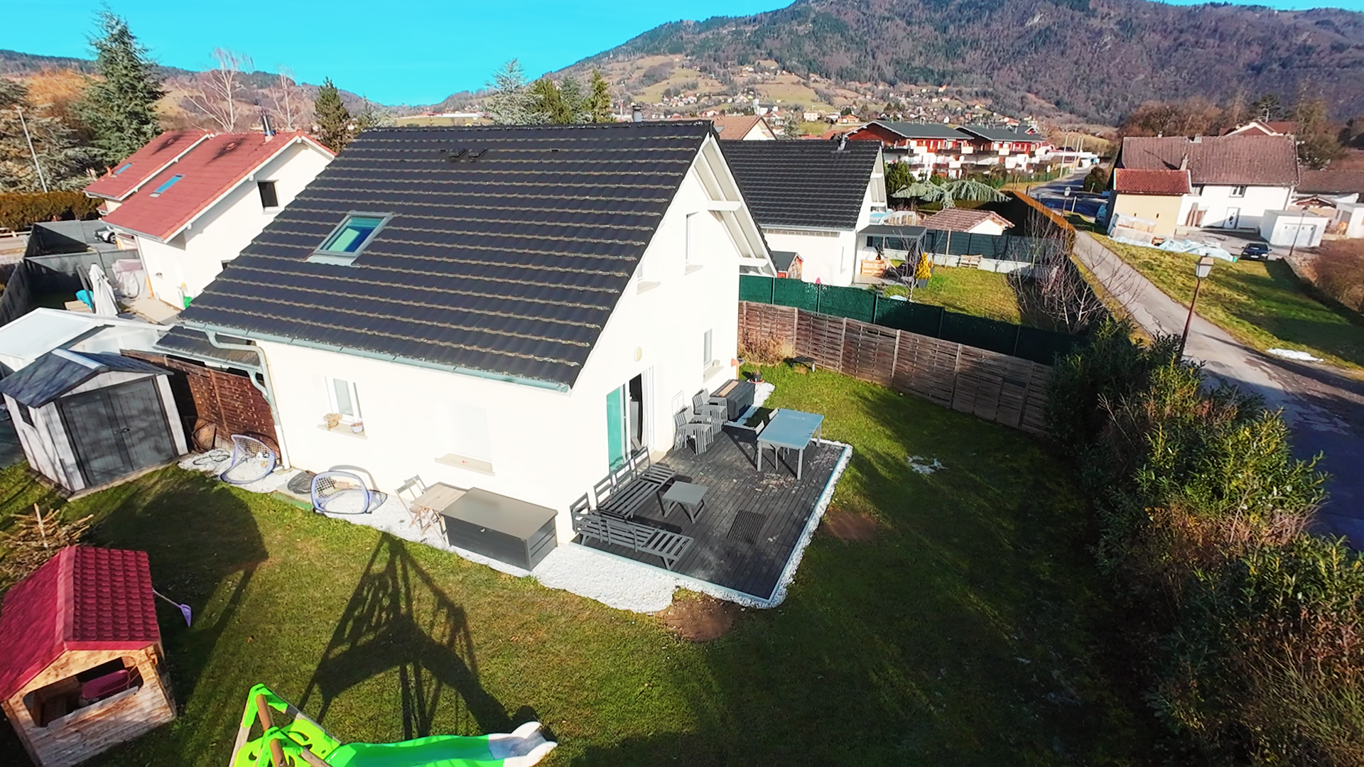Une maison blanche avec un toit en tuiles noires, une terrasse en bois avec des meubles, un jardin verdoyant, et un terrain de jeu pour enfants avec une glissière verte, dans un environnement résidentiel avec des montagnes en arrière-plan.