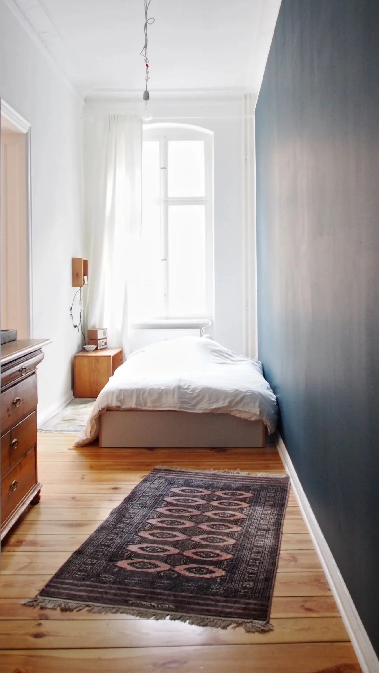 Ein Schlafzimmer mit Holzboden, einem weißen Bett, dunkler Wand auf der rechten Seite, einer kleinen Nachttischlampe, einem Fenster mit weißen Vorhängen, und einem bunten Teppich auf dem Boden.