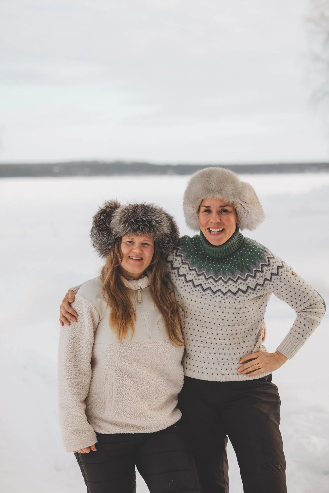 Kuva, jossa Kristiina ja Jaana eli naiset Tunne Lapin taustalla seisovat kädet toistensa ympärillä, suuret karvalakit päässä, ja hymyilevät kameralle.