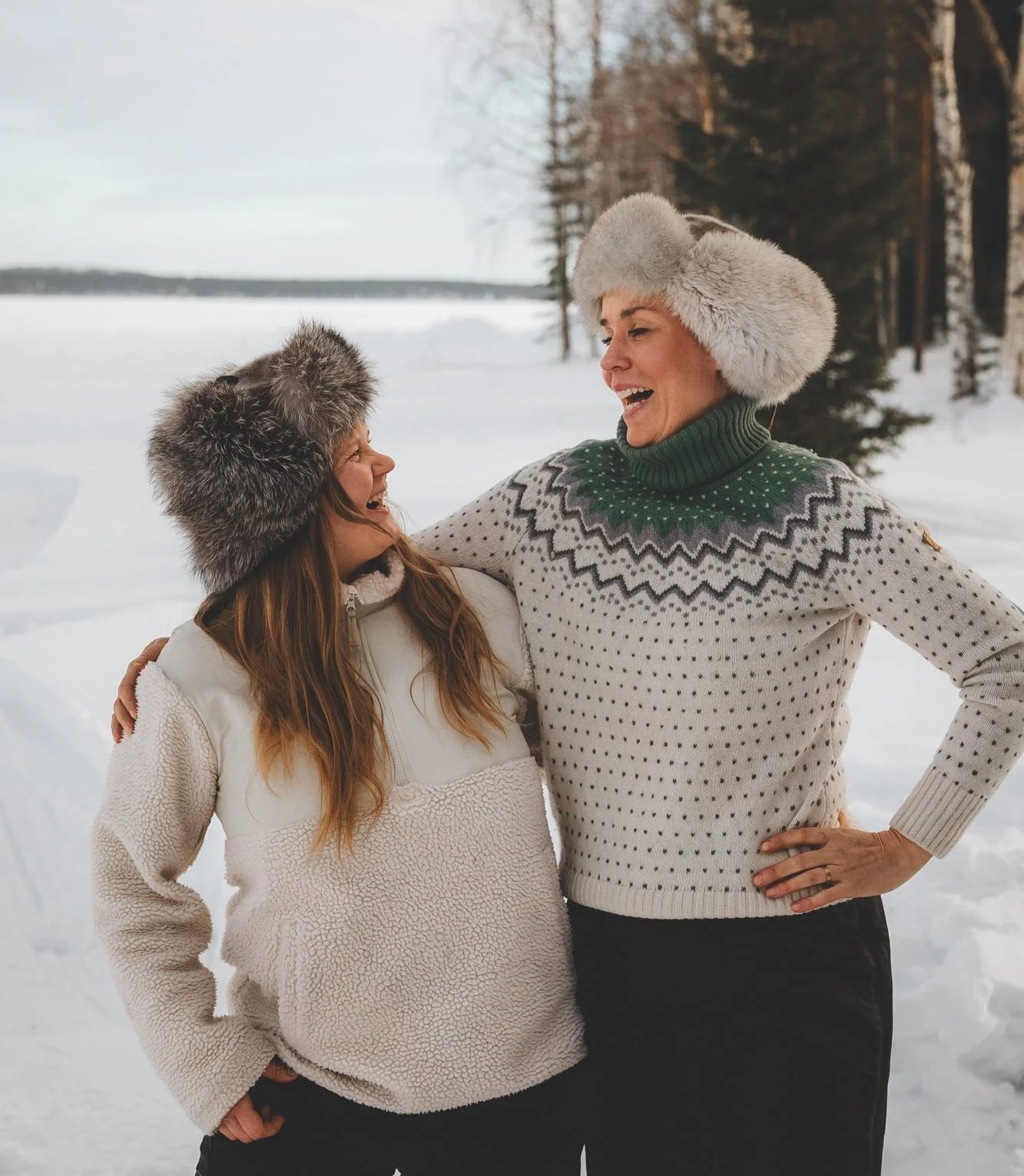 Kuva, jossa on luminen maisema, ja kaksi naista karvalakit päässä kädet toistensa ympärillä katsovat toisiinsa nauraen.