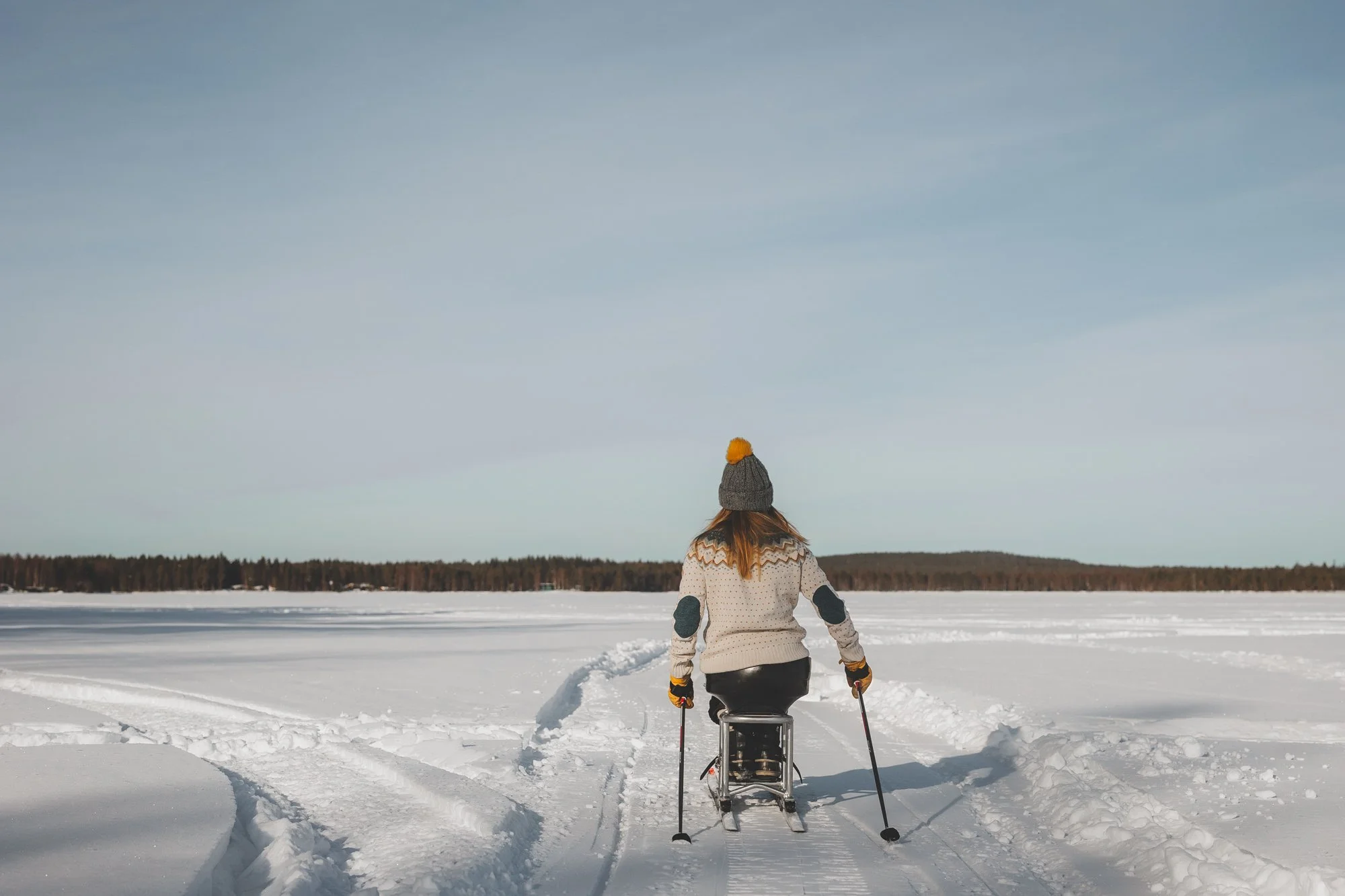 Nainen hiihtää suksipyörätuolilla, kuvattuna takaa.