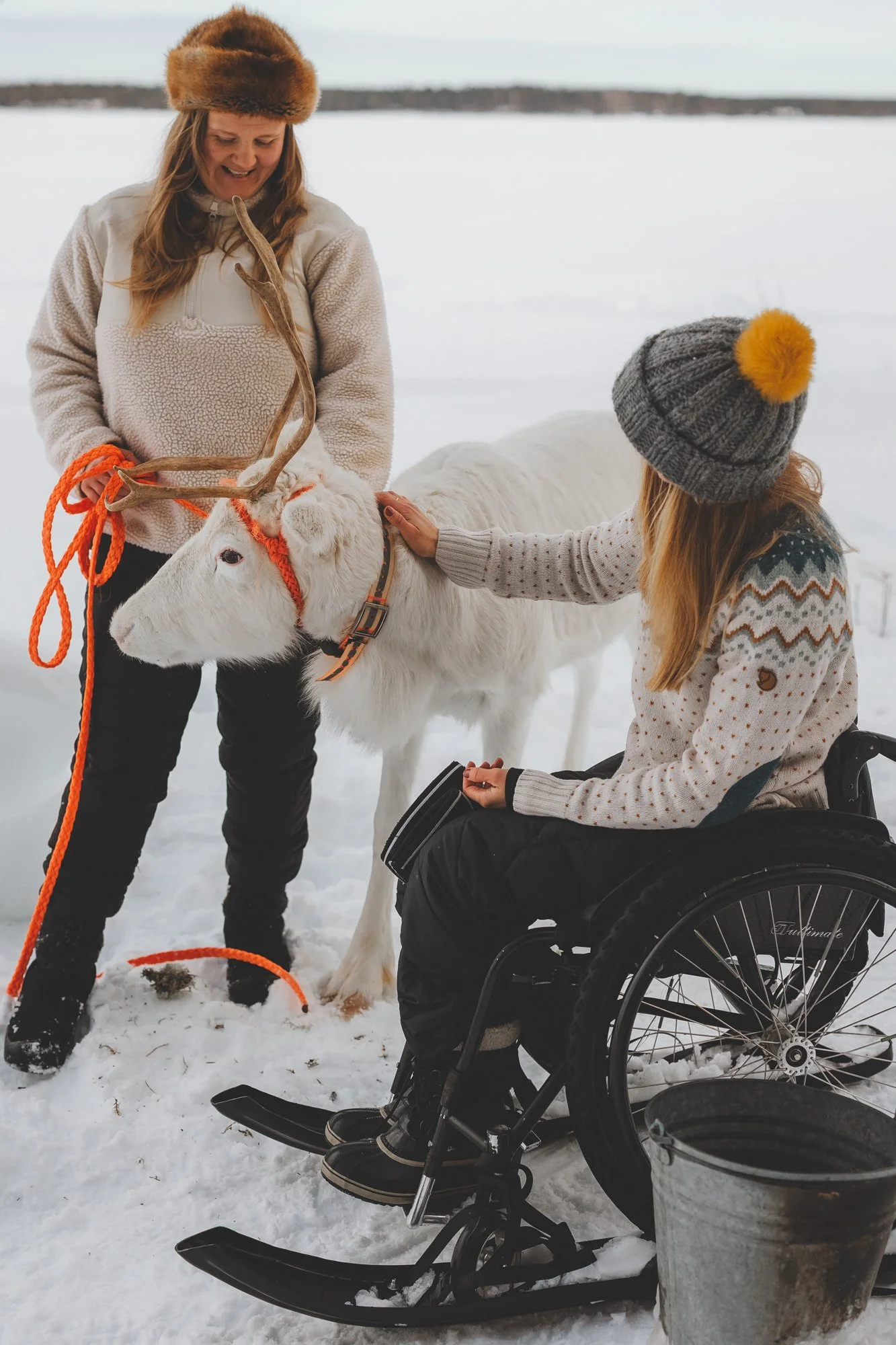 Kaksi naista järven jäällä. Toinen seisoo pitäen kiinni valkoisen poron talutusnarusta. Toinen istuu pyörätuolissa, jossa on sukset, ja silittää poroa.