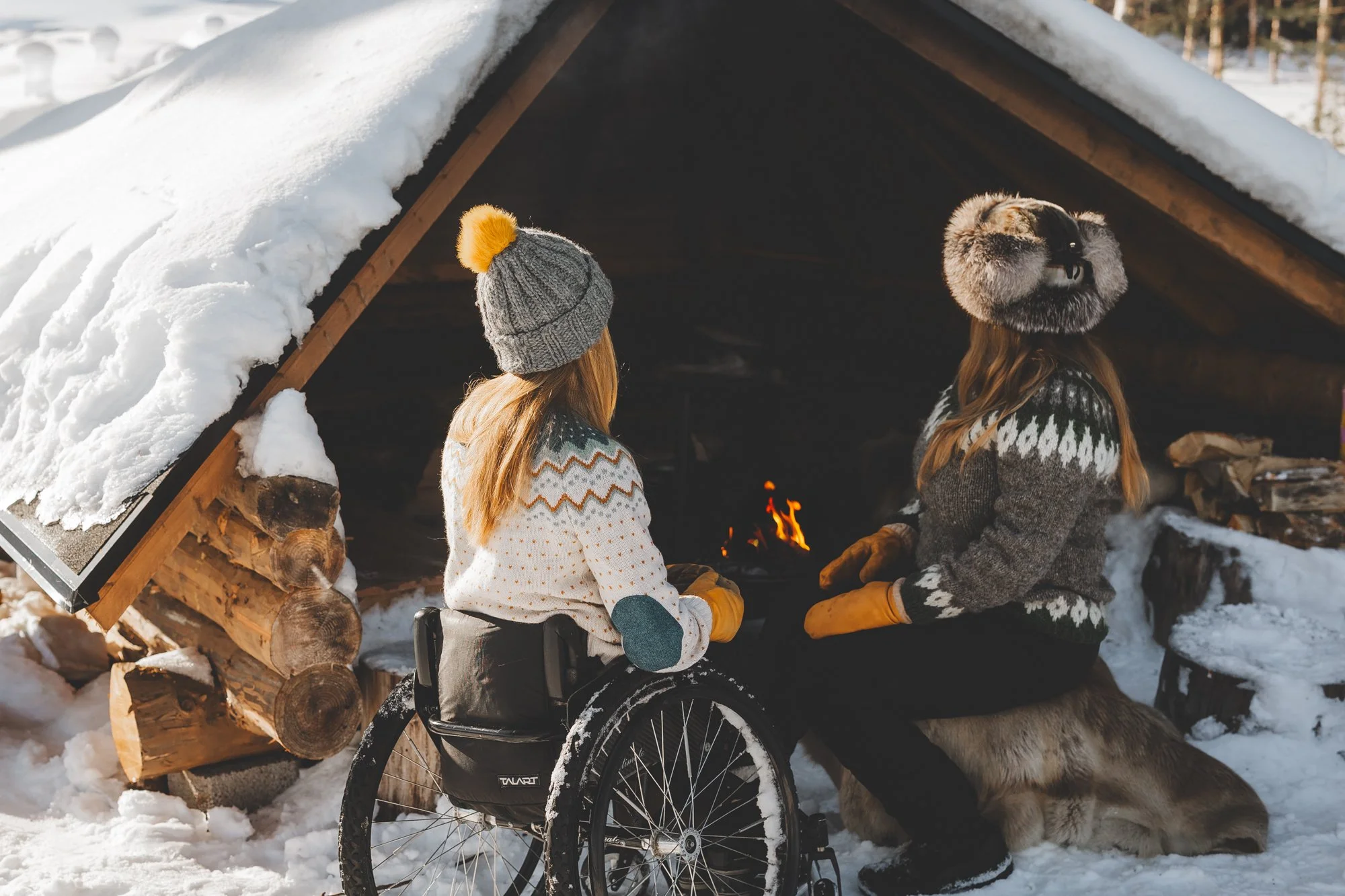Kaksi naista istuu laavun edessä ja nuotiossa palaa tuli. Toinen nainen istuu pyörätuolissa ja toinen poron taljan päällä.