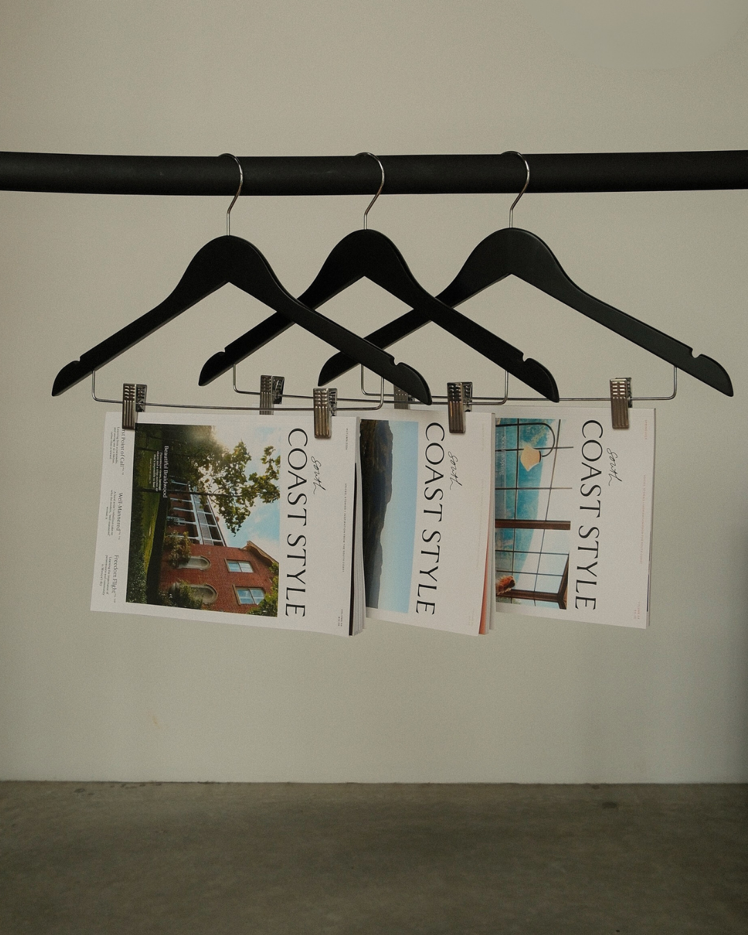 Three magazines titled 'Coast Style' hanging on black hangers from a black clothes rod, with the magazines featuring images of outdoor scenes and interiors.
