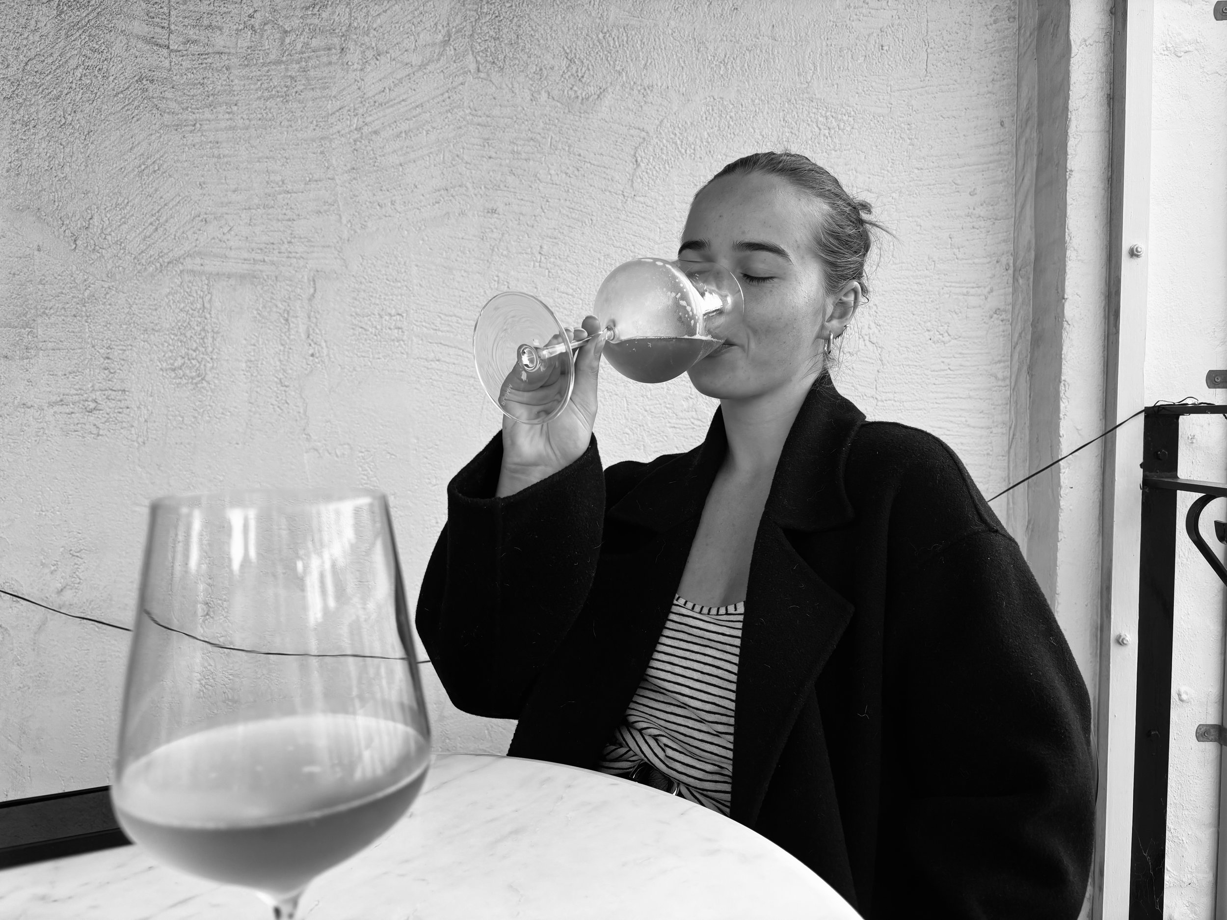 A woman with her eyes closed is drinking red wine from a glass at a table. The photo is in black and white.