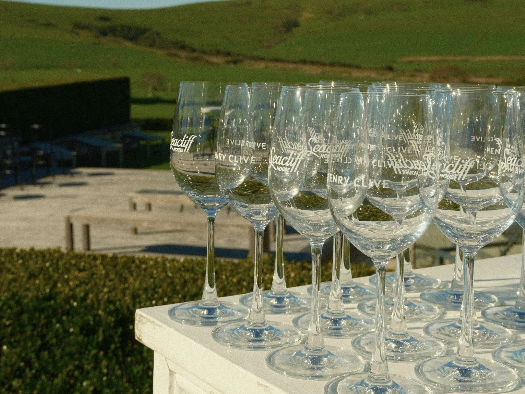 Multiple clear wine glasses with the words 'Lea cliff' and 'Henry Clive' printed on them, arranged on a white surface outdoors, with a green hillside in the background.