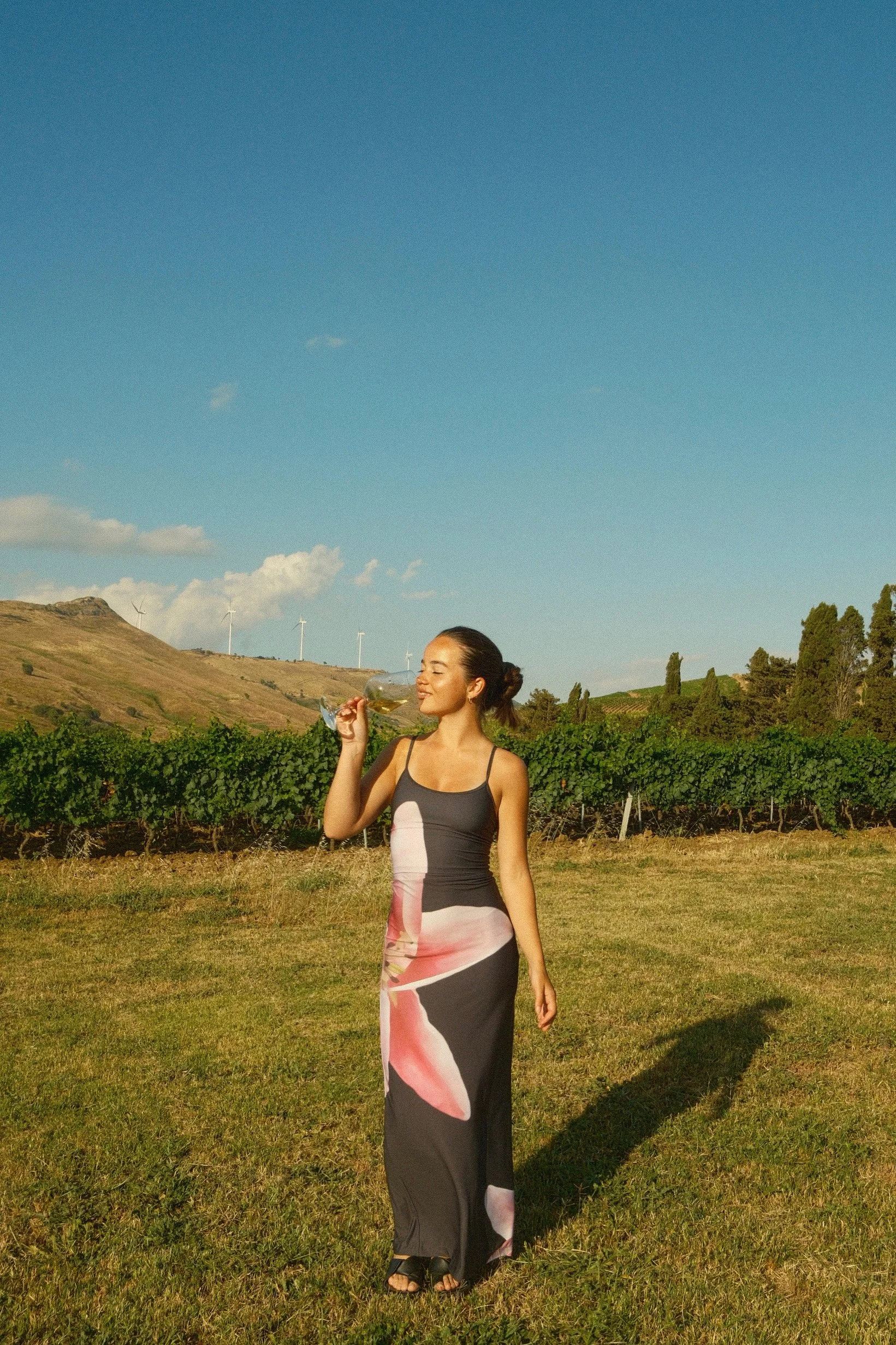 A woman in a colorful, sleeveless dress stands outdoors on a grassy field, holding a glass of white wine, with rolling hills, wind turbines, and a blue sky with clouds in the background.