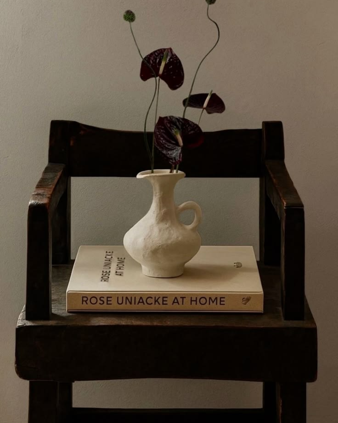 A white ceramic vase with a handle and a narrow opening, containing dark purple anthurium flowers with long, curly stems, placed on a closed book titled 'Rose Uniacke At Home' on a wooden chair, against a plain wall.