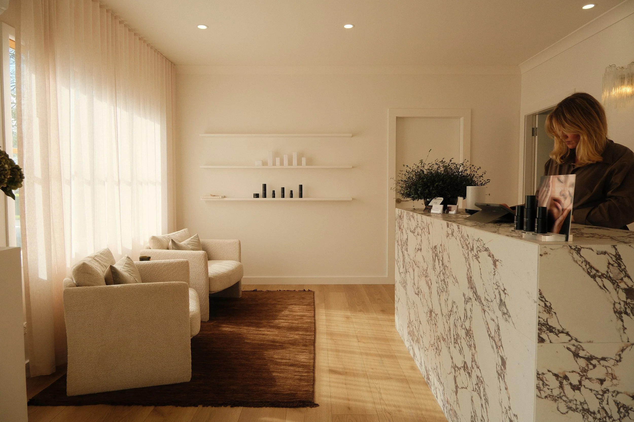 Interior of a modern, minimalistic lobby or reception area with beige armchairs, a brown rug, a marble reception desk, and an off-white wall with floating shelves and decorative objects. A woman stands behind the desk using a phone.