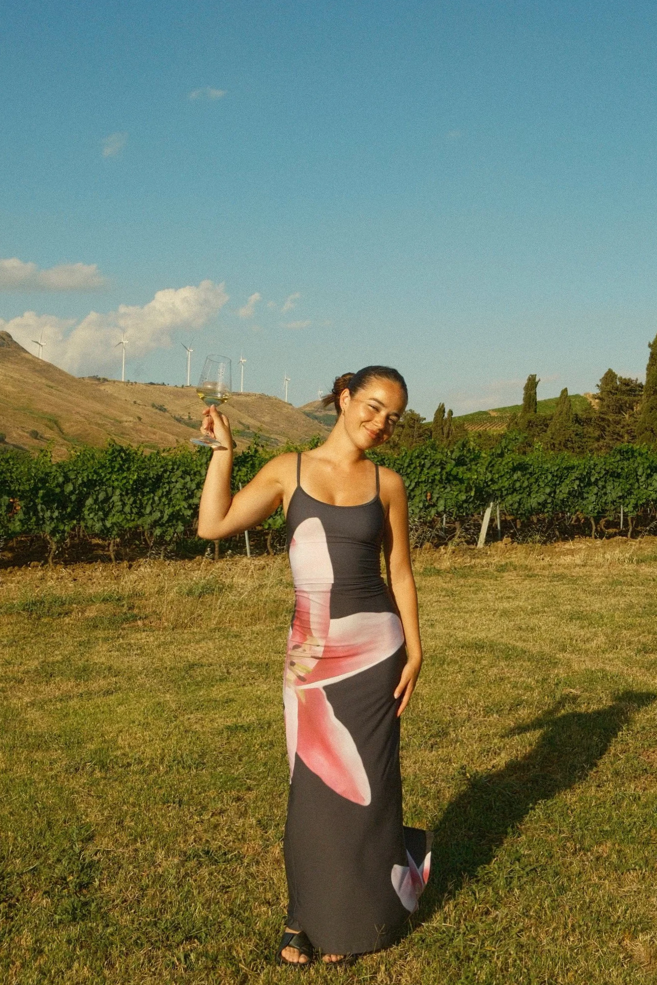 A woman in a colorful dress standing in a vineyard holding a glass of wine, with wind turbines and hills in the background on a sunny day.