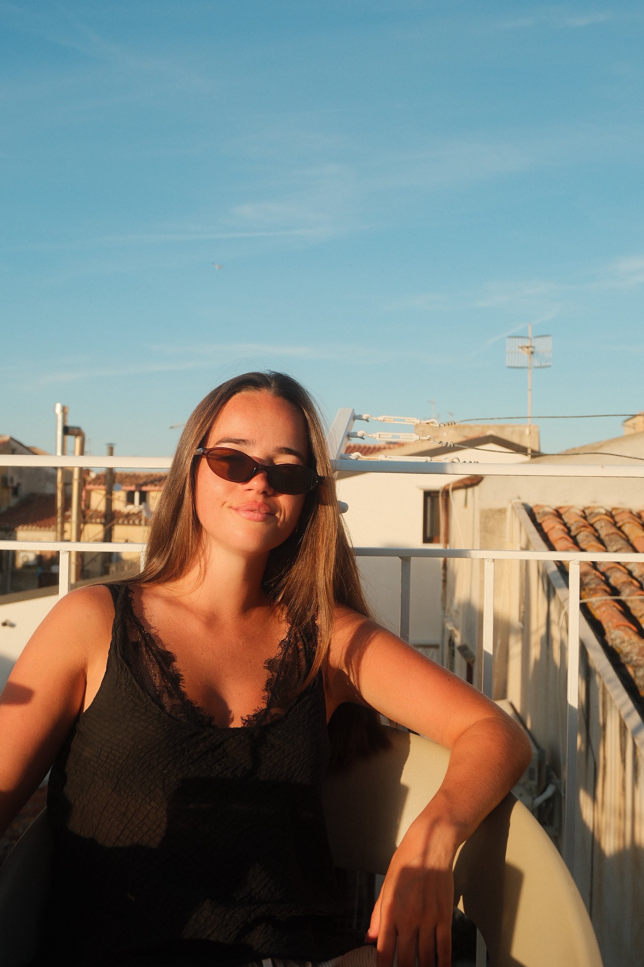 A woman with long hair wearing sunglasses and a black sleeveless top, sitting on a balcony with rooftops and a clear blue sky in the background, taking a selfie.