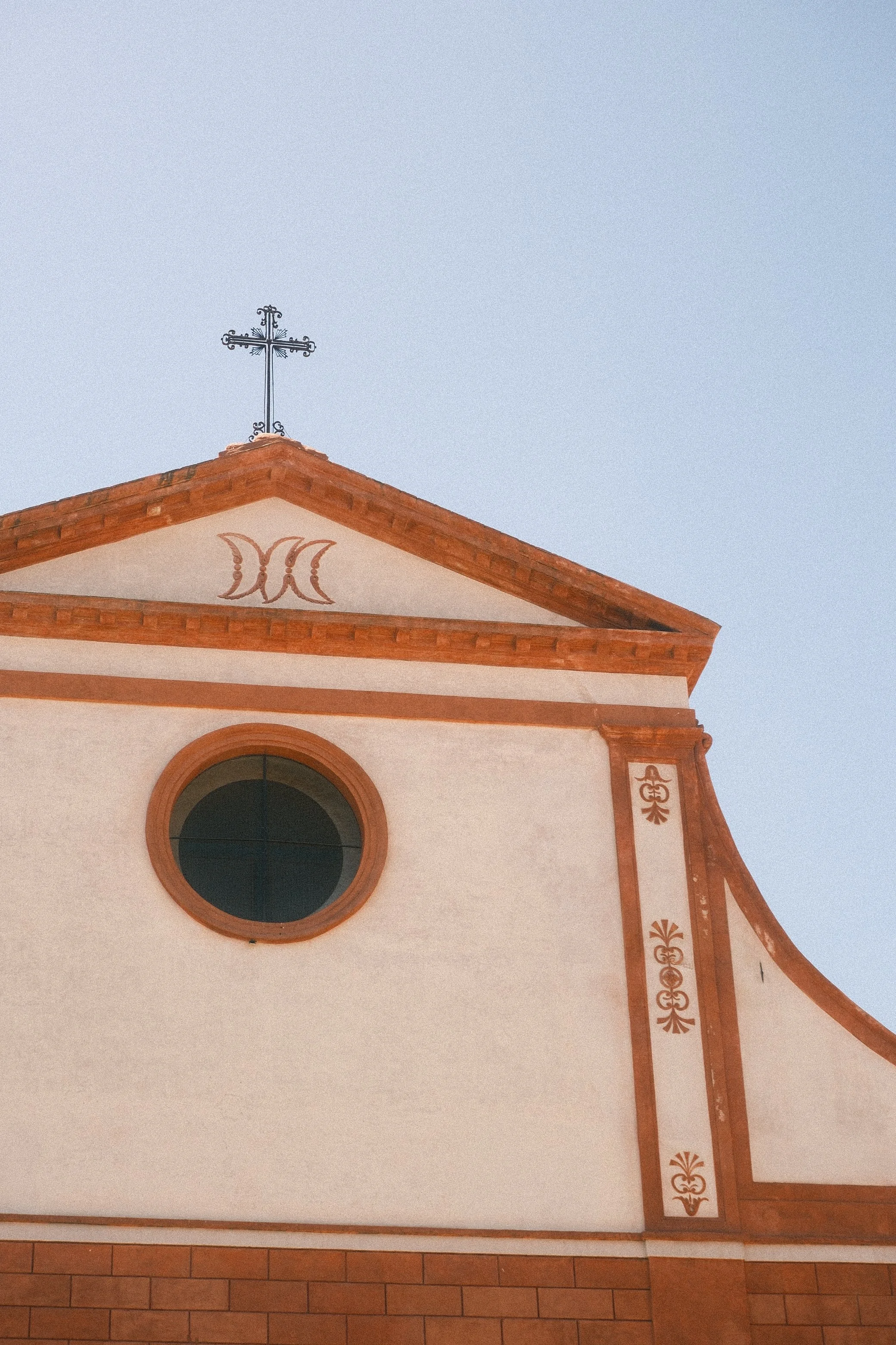 Photo of a church facade with a triangular roof, a round window, and decorative elements, topped with a metal cross against a clear sky.