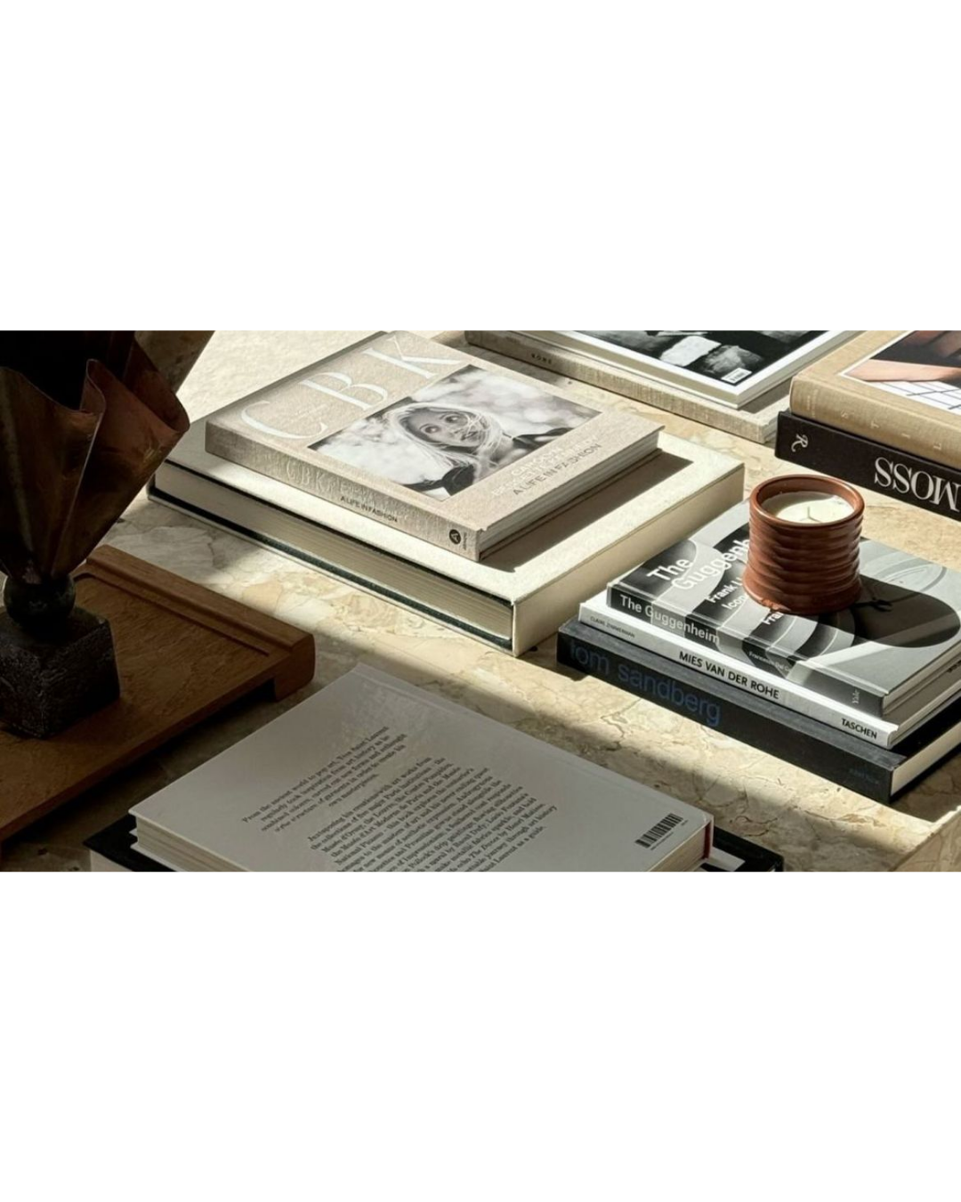 A marble tabletop displays various art and design books, a small potted candle, and a wooden panel. The books include topics on portraiture, designers, and artists, with some stacked and others spread out, illuminated by sunlight.
