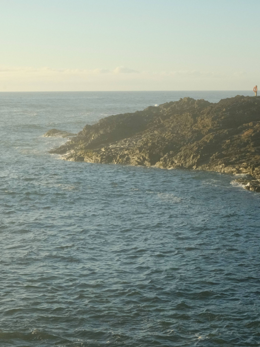 Ocean waves hitting rocky shoreline with a distant horizon and a small navigation marker in the background.
