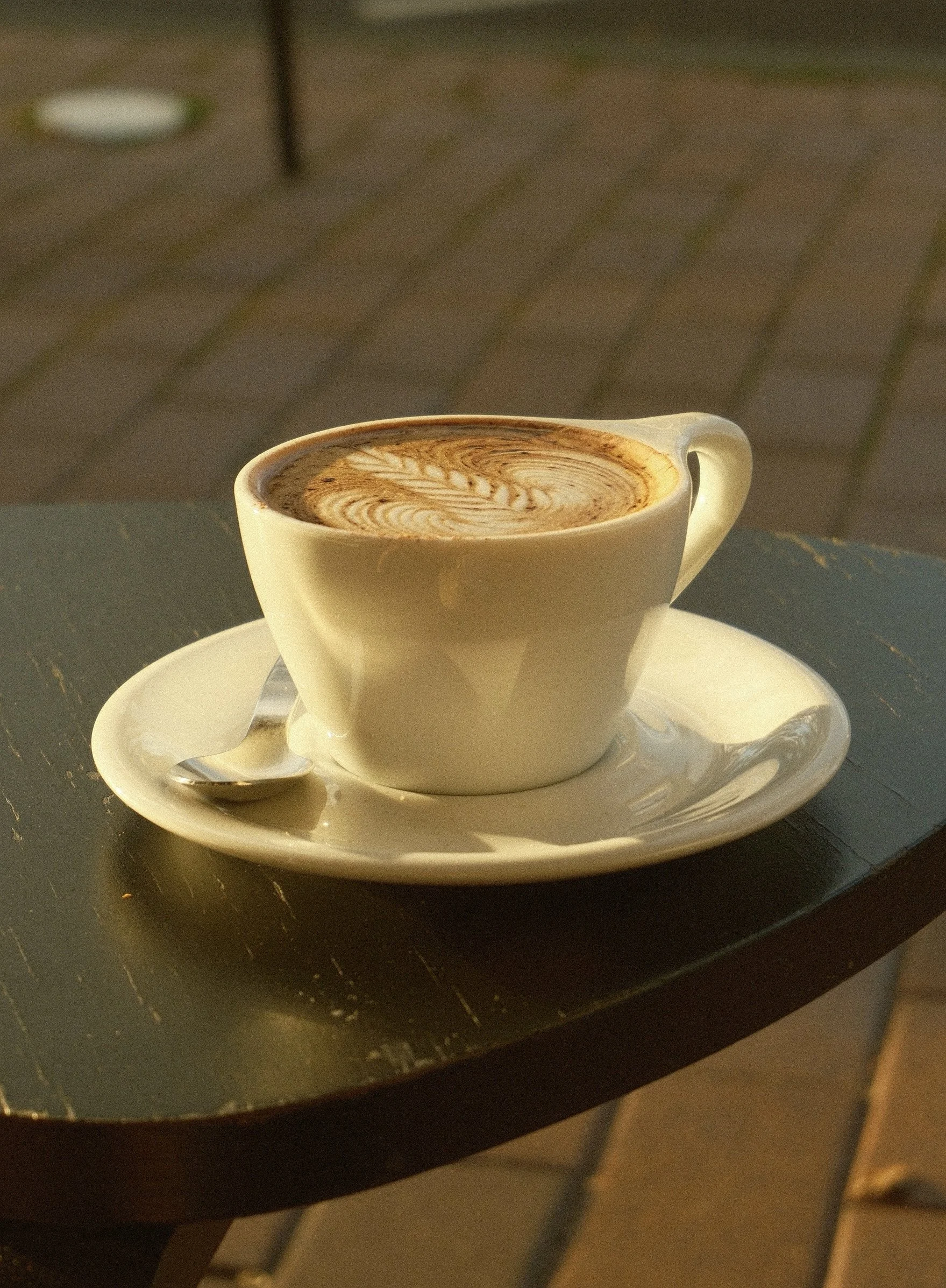 A white cup of hot chocolate with latte art on top, served on a small white saucer with a small silver spoon, placed on a dark table.