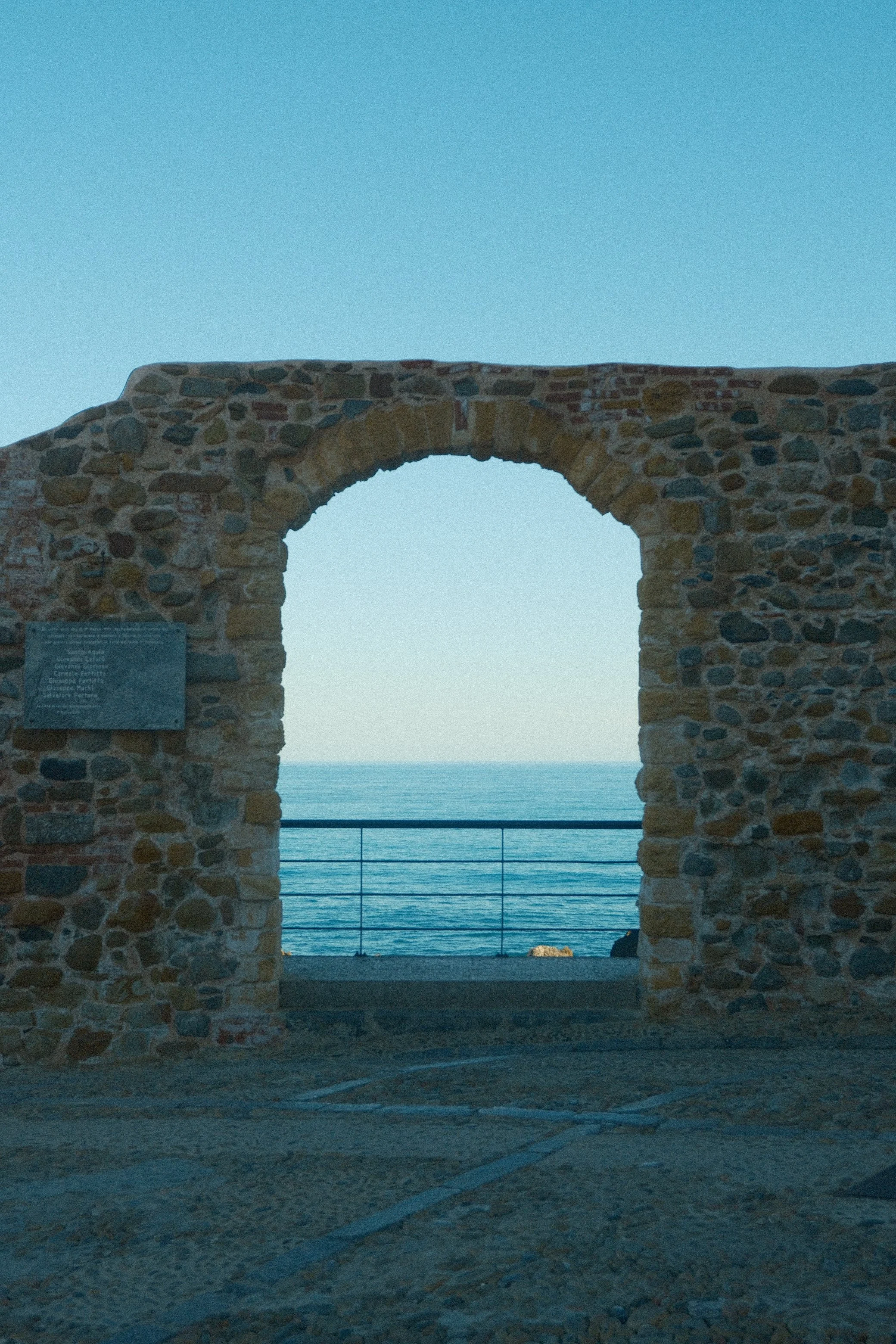 A stone archway overlooking a body of water, with a railing, against a clear blue sky.