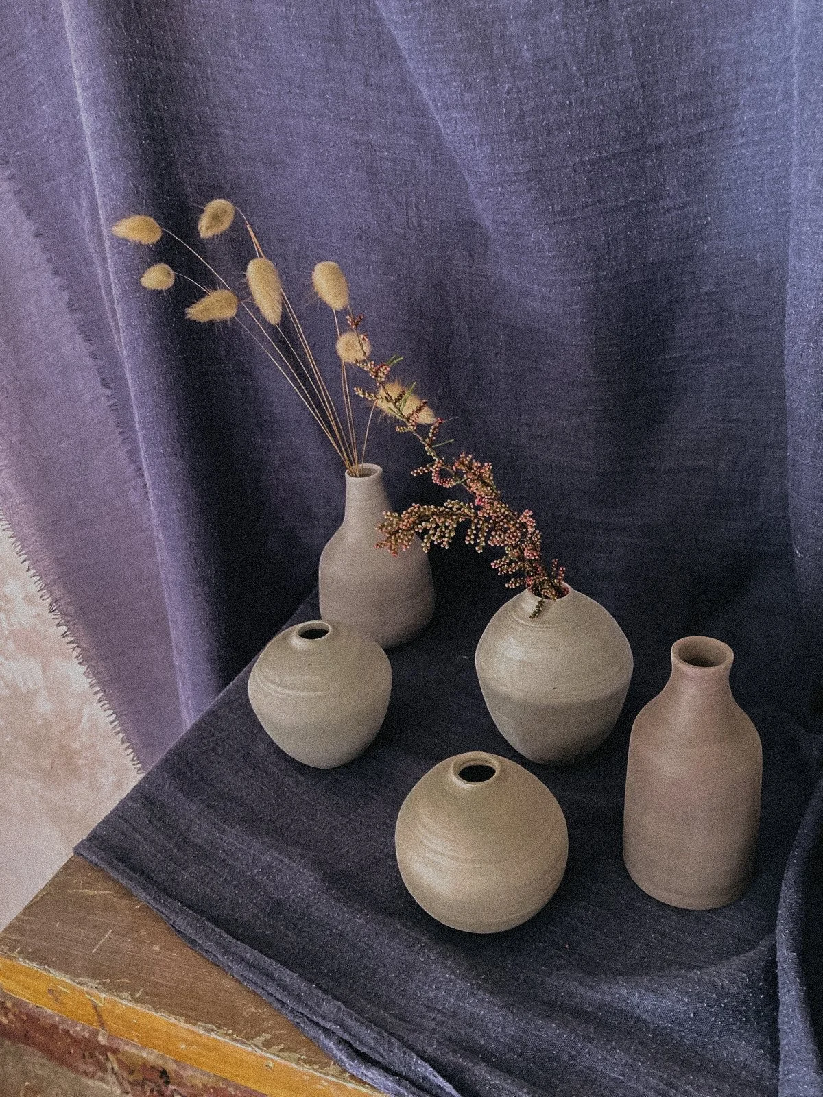 Cinq vases en terre cuite beige contenant des fleurs séchées, placés sur une surface avec un tissu gris foncé en arrière-plan.