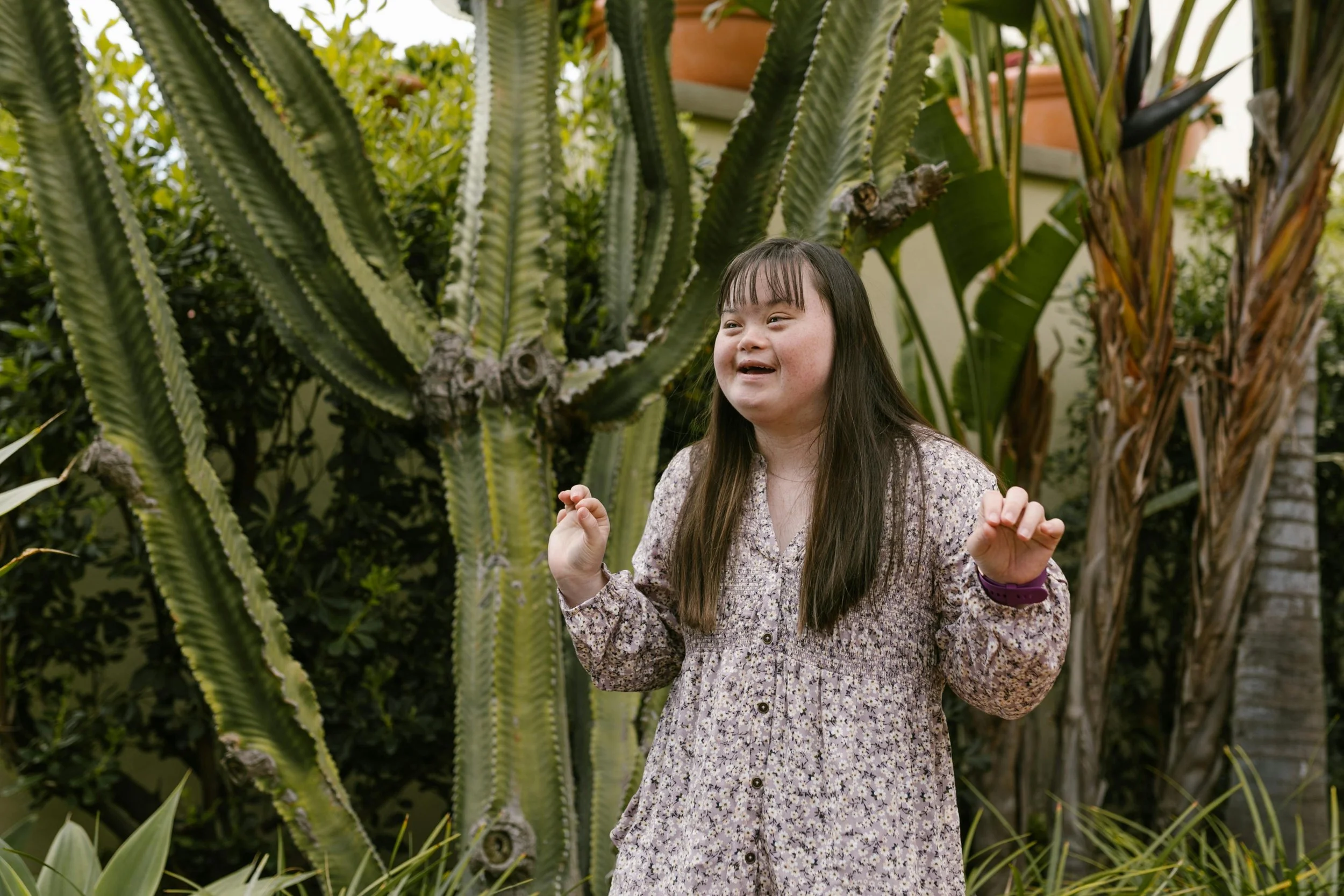 A young girl with Down syndrome standing outdoors in front of large cactus and other tropical plants, smiling and gesturing with her hands.