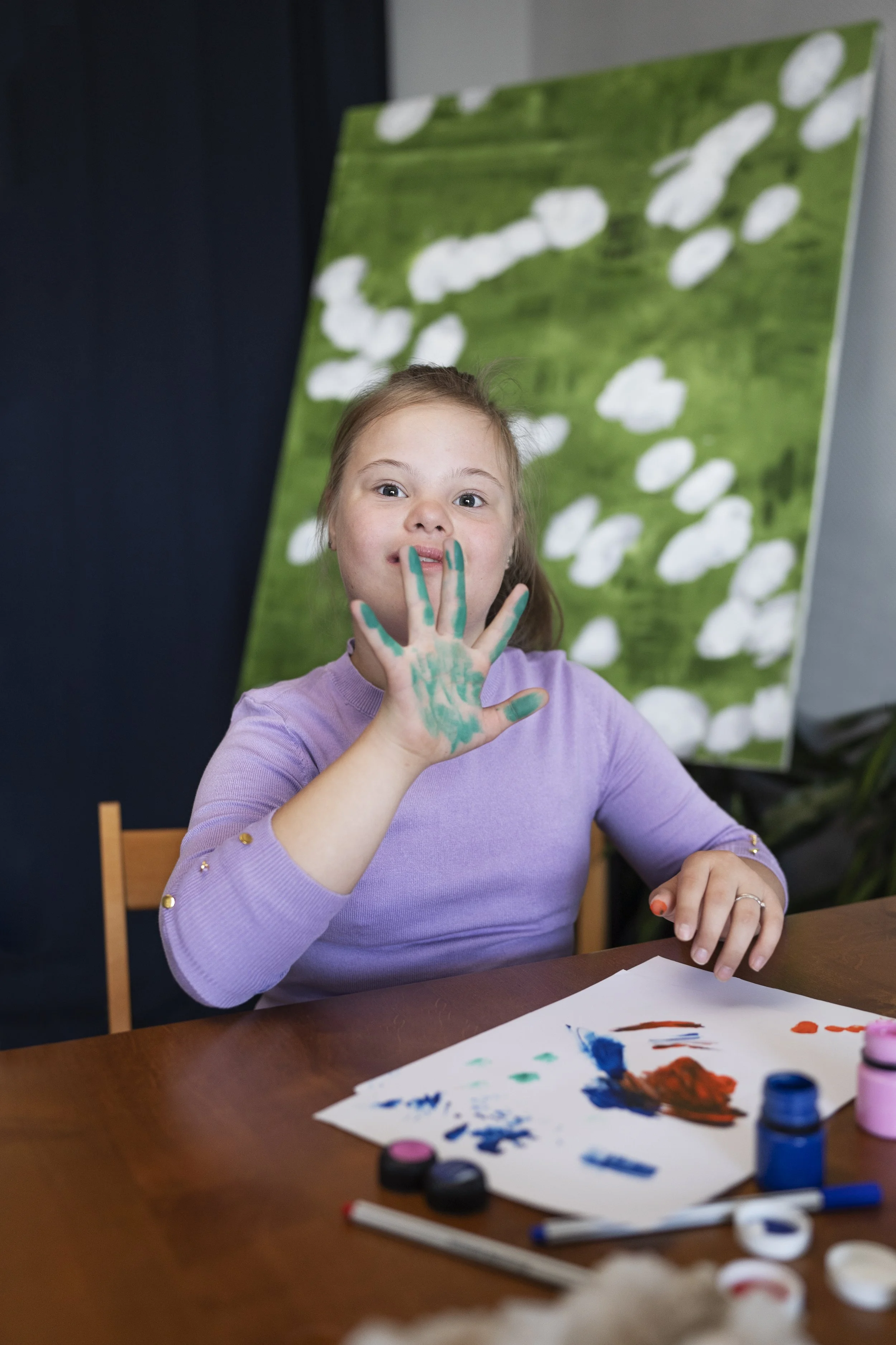 A young girl with paint on her hand and face, sitting at a table with paint supplies, holding up her hand to show the paint.