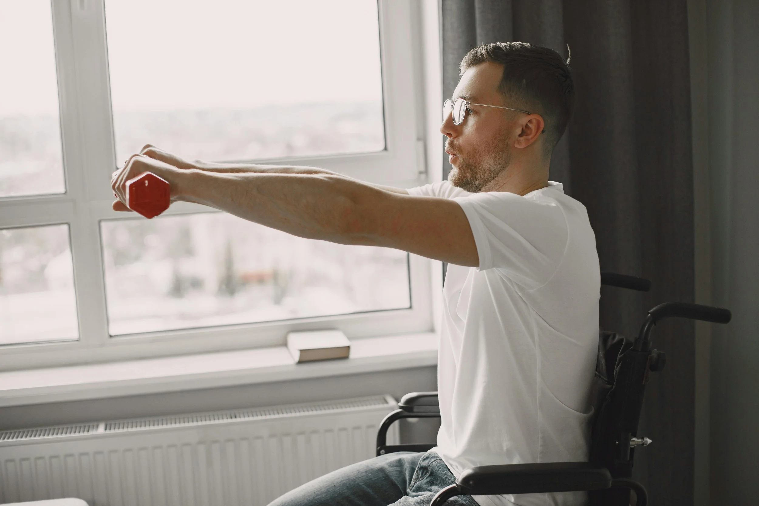 A man in a wheelchair doing arm exercises with a dumbbell in front of a window.