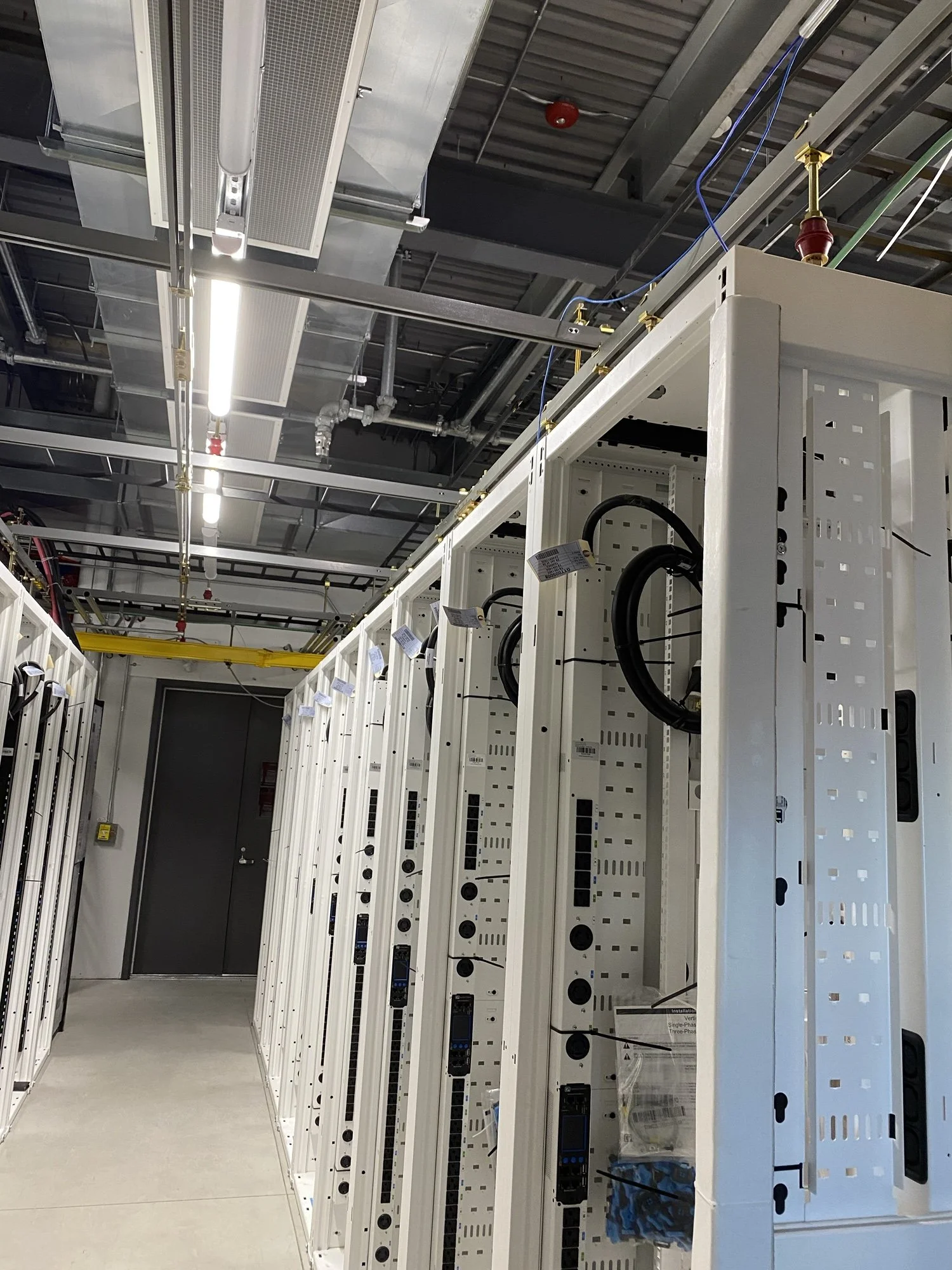 Server racks and network equipment inside a data center with overhead lighting and exposed piping and wiring.