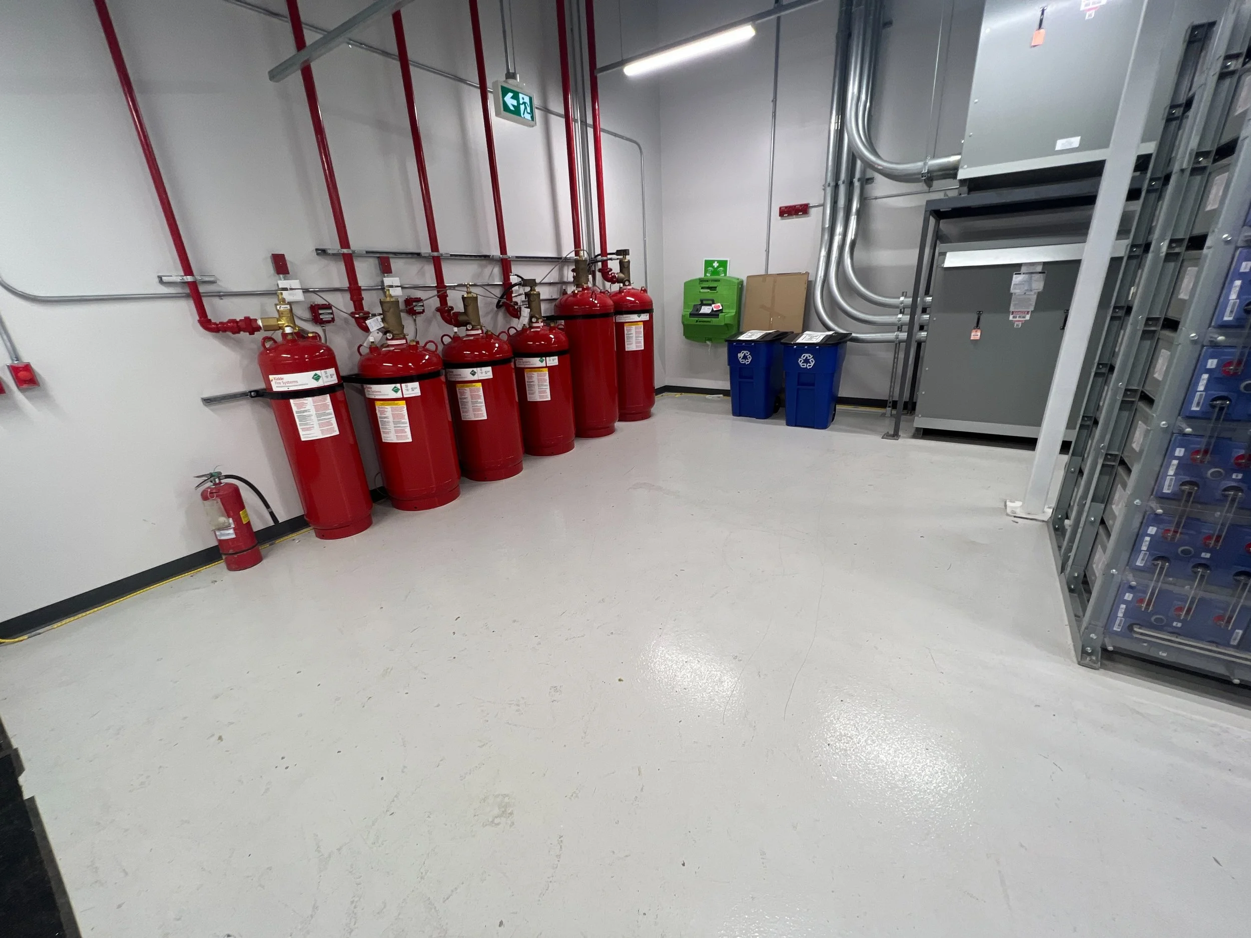 A fire safety room with six red fire extinguishers on the wall, a smaller fire extinguisher on the floor, green emergency equipment, recycling bins, and electrical panels.