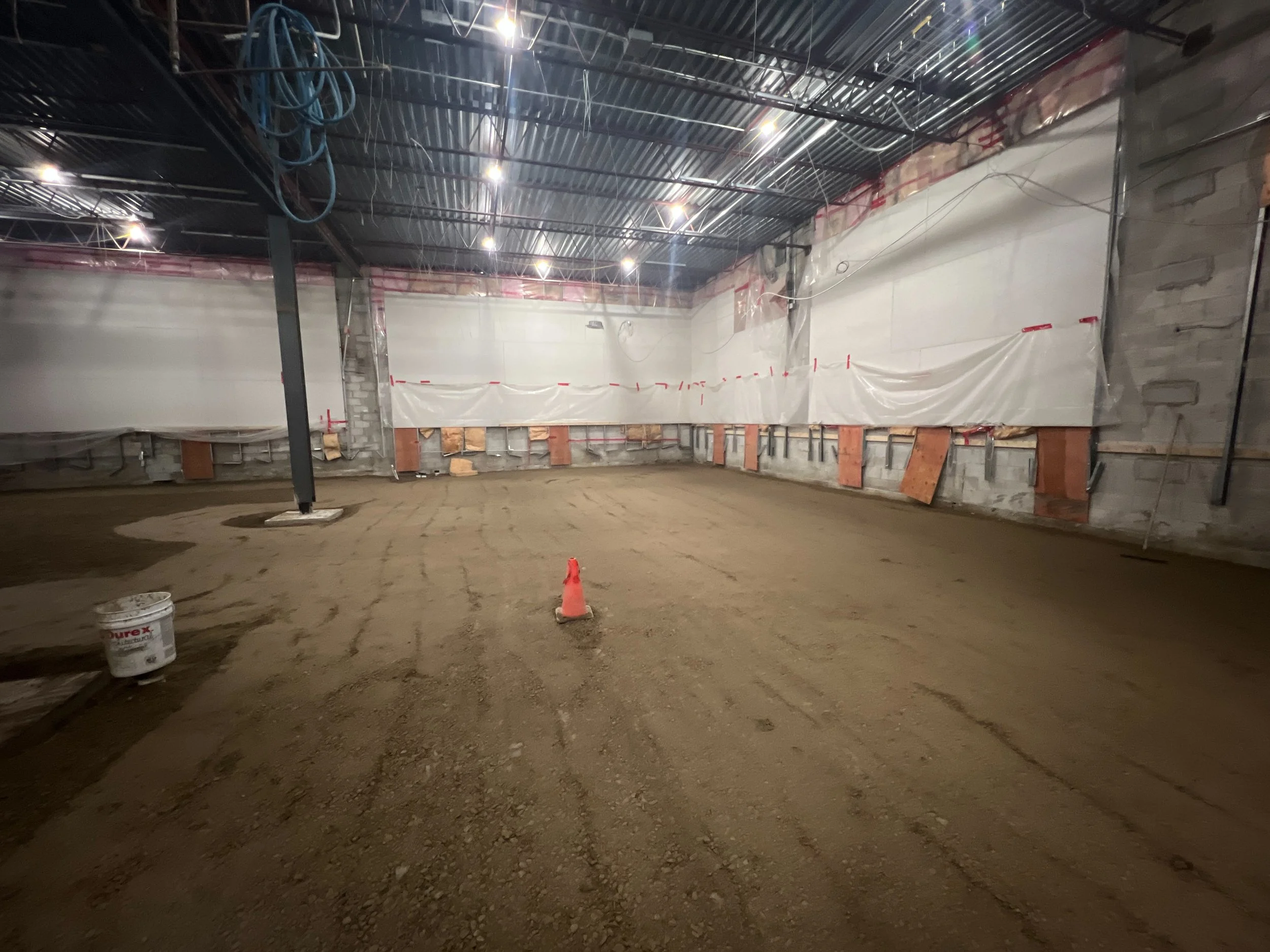 An indoor construction site with an unfinished interior, featuring a dirt floor, plastic sheeting on the walls, exposed ceiling with wiring, and construction materials and cones.